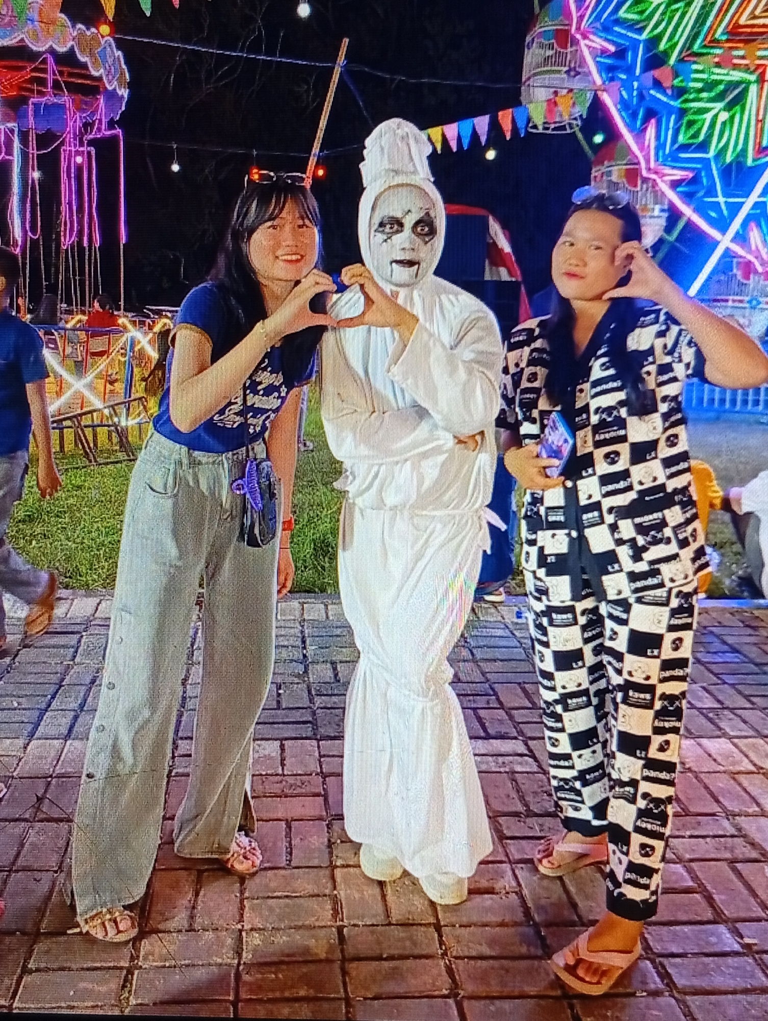 In Lhokseumawe, Indonesia, three individuals pose for a photograph at an amusement park at night. On the left, a young woman wearing a blue t-shirt and grey pants smiles at the camera, forming a heart shape with her hands. To her right stands a person in a white ghost costume with face paint, also making a heart shape with their hands. On the far right, another young woman in a black and white panda-patterned pajama set stands with her hand on her cheek. In the background, a brightly lit Ferris wheel and carousel are visible, along with other park attractions and a crowd of people. The ground is paved with interlocking bricks.
