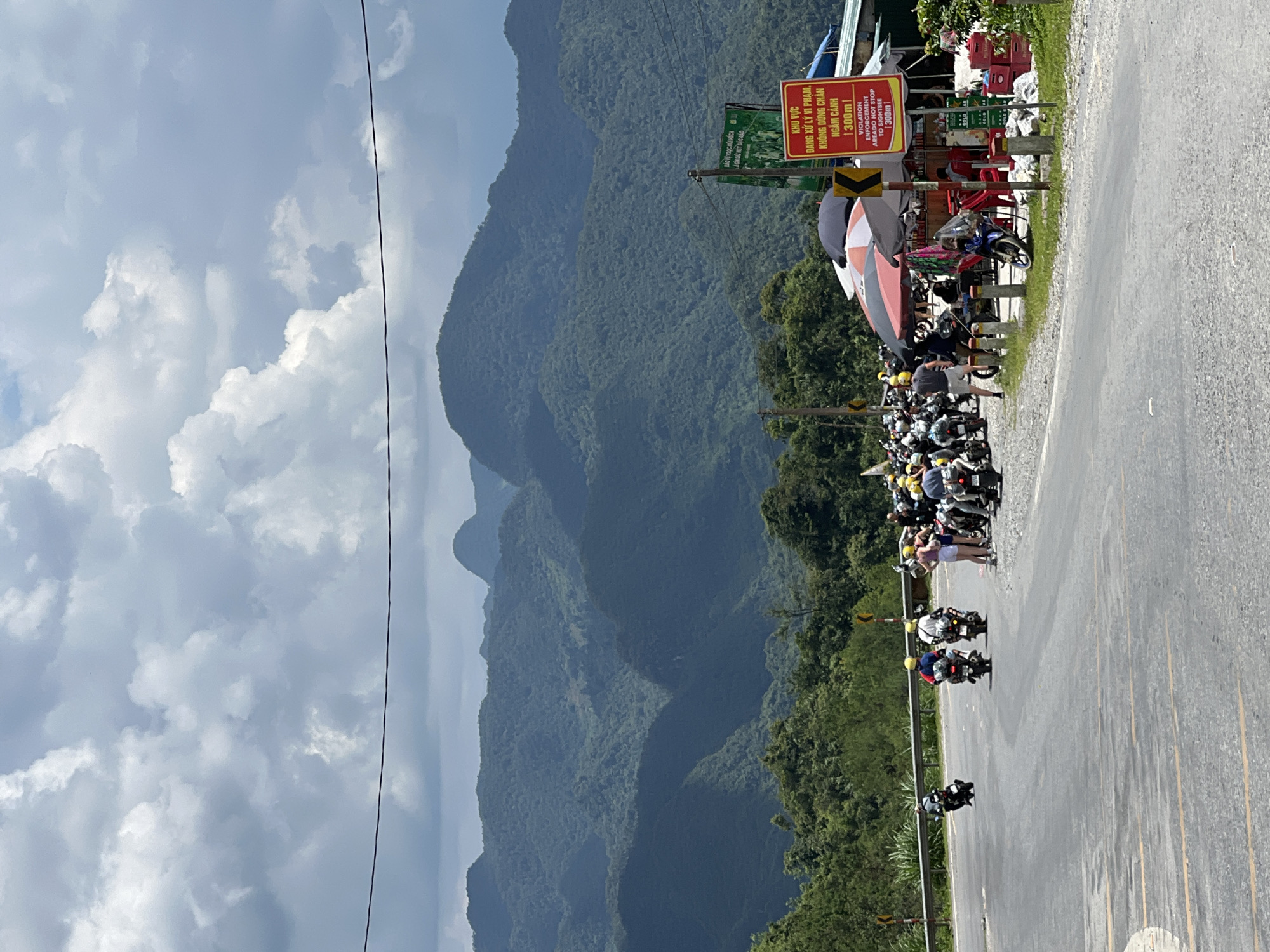 A group of motorcyclists is gathered on the side of a winding mountain road in Quản Bạ Commune, Vietnam, with a backdrop of lush, verdant mountains under a cloudy sky. The road is paved with asphalt and marked with yellow lines. In the foreground, several motorcycles are parked, and some individuals are standing or sitting near a small roadside stall with umbrellas. The stall displays various items, including drinks and red crates. A red and yellow sign on a pole reads "KHU VỰC DỪNG CHÂN NGẮM CẢNH" and "VIOLATION ENFORCEMENT AREA TO STOP TO SIGHTSEE 300m". Further down the road, a few motorcyclists are in motion, navigating the curves. The mountains in the background are covered in dense forest and appear layered, with shadows cast by the clouds. A thin black wire is visible stretched across the upper portion of the image.