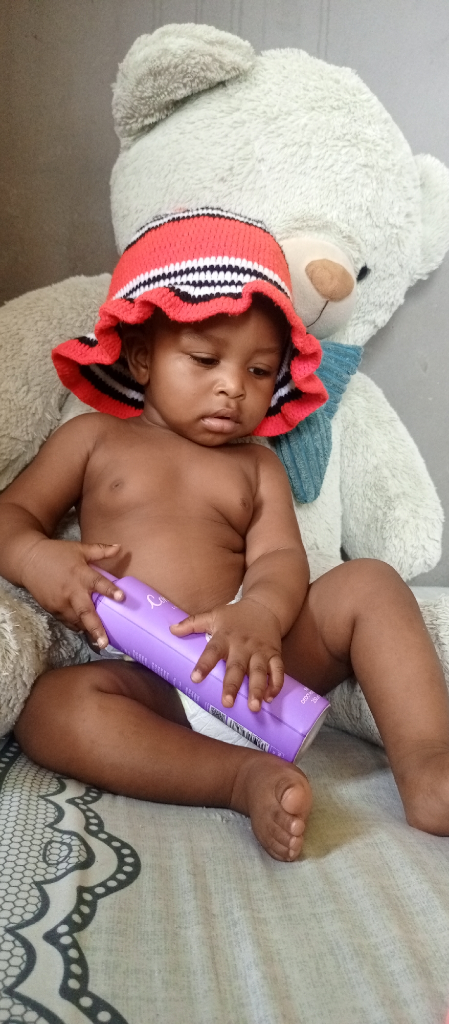 An overhead, eye-level shot shows a baby or toddler sitting indoors, leaning against a large plush teddy bear. The baby, with dark skin, is wearing only a white diaper and a red, white, and black striped knitted sun hat with a ruffled brim. Their expression is focused and contemplative as they look down at a long, rectangular, purple bottle or container held in both hands.
The large, light-colored (likely off-white or light grey) teddy bear sits directly behind the baby, its head and torso visible. It has a light brown snout and nose, and a ribbed light blue scarf tied around its neck. The baby's left arm rests on the bear's fluffy body.
The purple bottle held by the baby has some white text. The word "Care" or a similar cursive word is faintly visible near the top left side. Towards the bottom right, "DEOXOO" and "250ml" are readable, along with a barcode.
The setting appears to be a domestic indoor space, possibly on a bed or cushioned surface covered by a light grey patterned sheet with dark grey and white geometric designs. The background is a plain, light-colored wall or surface. The lighting is soft and even, suggesting daytime or well-lit interior conditions in Keffi, Nigeria.