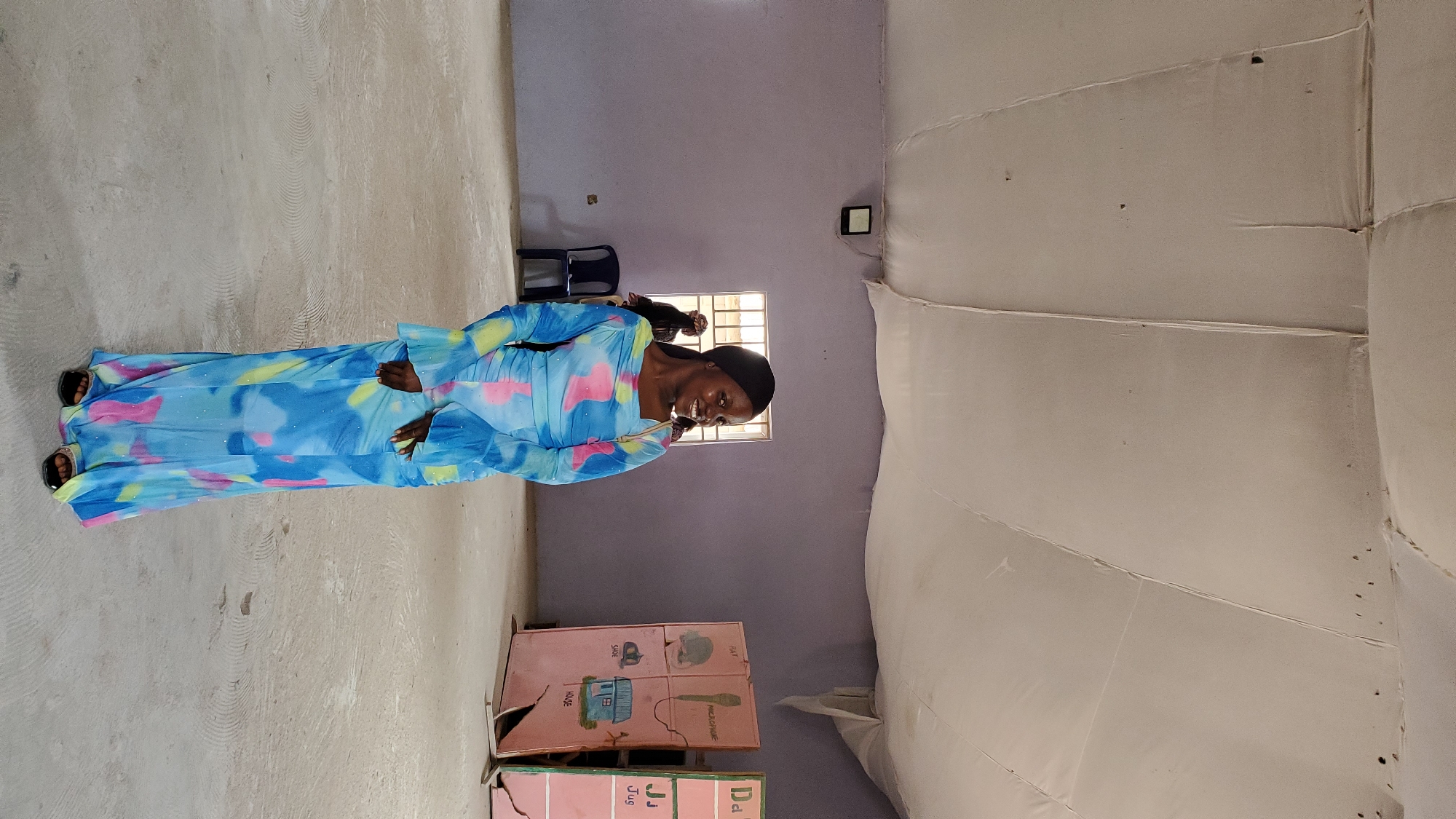 The image shows a young Black woman standing in an indoor space, likely a community hall or classroom, in Jalingo, Nigeria. She is wearing a long, flowing blue dress with a tie-dye pattern of pink, yellow, and blue. The dress has flared sleeves and is cinched at the waist. She is smiling and looking to her right.
The environment appears to be sparsely furnished. The walls are painted a light purple. A window with a grate is visible in the background, and behind it, two other individuals, their faces obscured, are standing. To the left of the window, a dark blue plastic chair is placed on the concrete floor. On the right side of the room, two large, pink display boards are leaning against the wall. One board has images and text that appear to be educational, with words like "RAT," "MICROPHONE," "SHOE," and "HOUSE" visible. The other board is partially obscured and also seems to contain educational content. The ceiling is made of white fabric stretched across beams, creating a somewhat uneven and draped appearance, possibly indicative of temporary or basic construction. The floor is a light gray concrete surface with visible circular patterns from what might be a polishing or cleaning process. A square light fixture is mounted on the upper left wall.