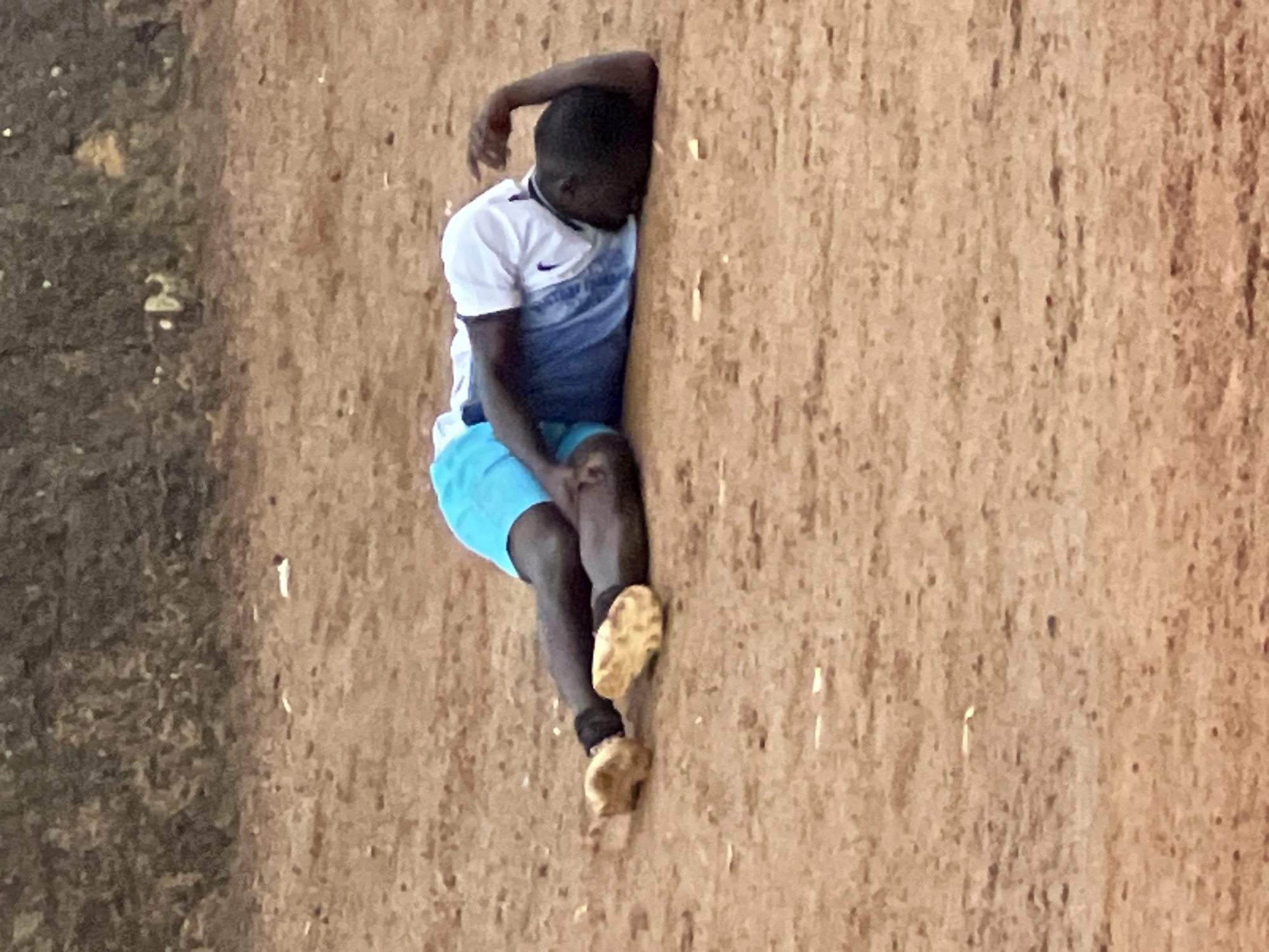 A young Black male is lying on the ground, on his right side, with his legs bent and feet pointing upwards. He is wearing blue shorts and a white t-shirt with blue accents and black trim. His right arm is bent at the elbow and resting on his chest, while his left arm is extended behind him, with his hand resting on the ground. He appears to be in distress, with his eyes closed and his head turned to the side. The ground is a reddish-brown dirt, and there is a textured, dark brown object in the background, possibly a wall or embankment. The scene is set in an outdoor environment, likely a dusty area, consistent with the dry, earthy conditions often found in parts of Jos, Nigeria. There are no other individuals or objects immediately visible that would provide further context to the situation.