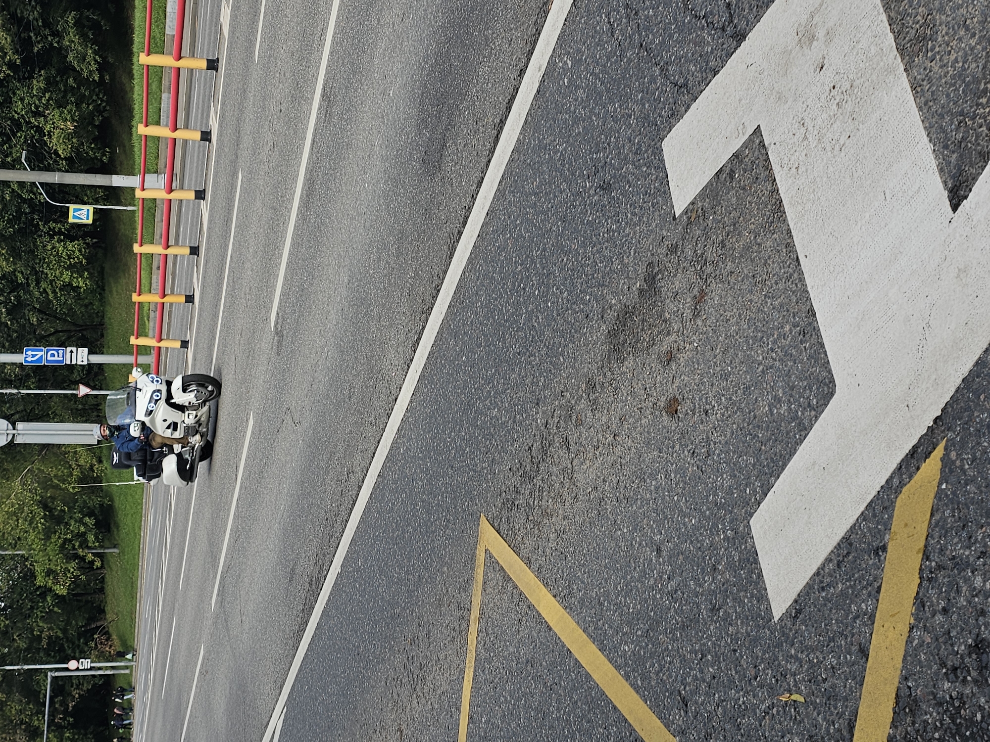 A white Honda Gold Wing motorcycle with a police officer rider is traveling on a multi-lane road in Moscow, Russia. The motorcycle is white with chrome accents and has a large trunk. The officer is wearing a blue jacket, brown pants, a helmet, and a face mask. The road is asphalt and has white lane markings. A yellow and red metal guardrail separates the road from a grassy embankment. There are trees and bushes on the embankment. Traffic signs are visible in the background, including a blue parking sign and a yellow triangle sign. The scene appears to be on a slightly overcast day.