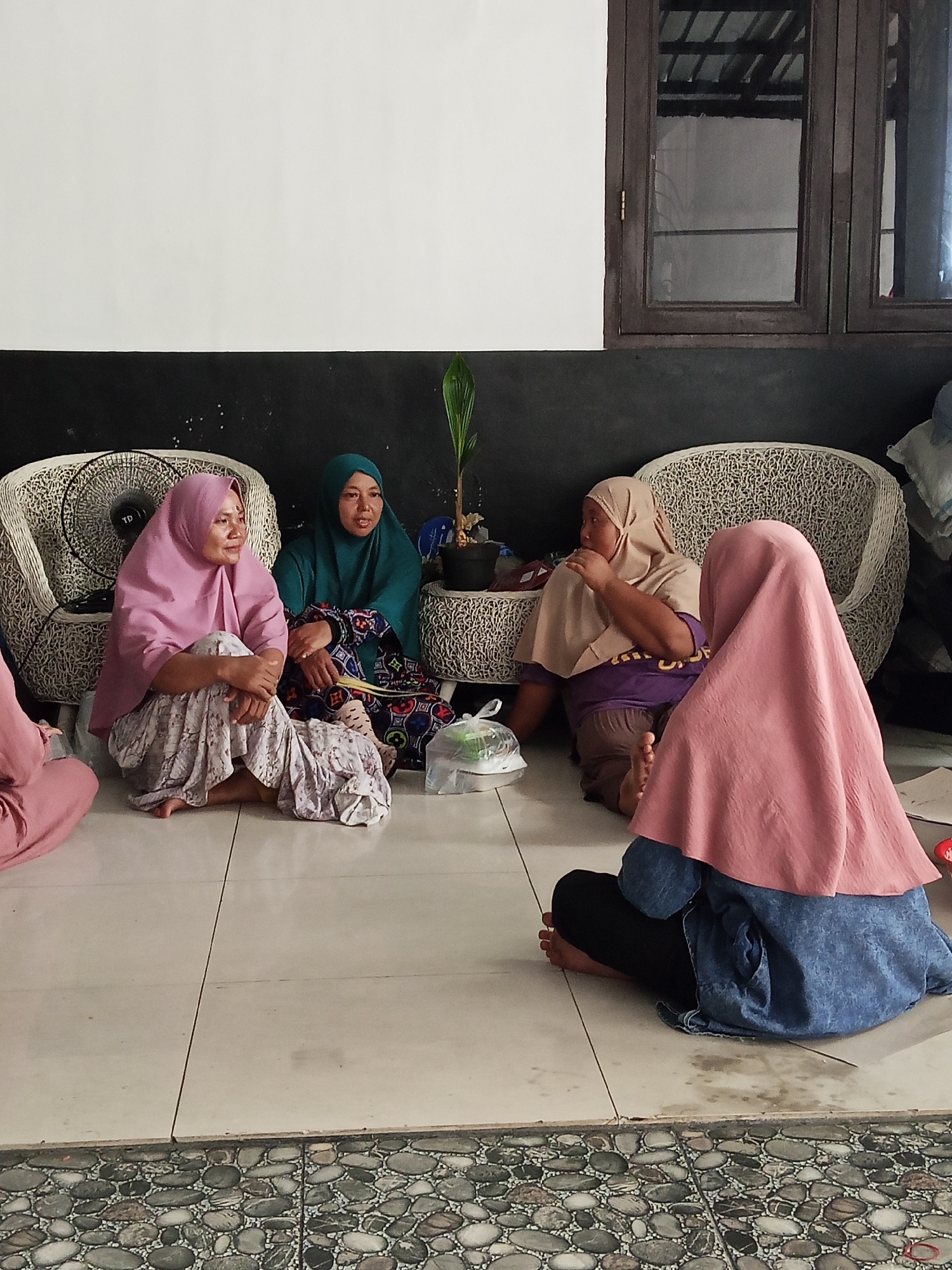 Four women are seated on the floor in what appears to be an outdoor or semi-outdoor covered area with a tiled floor in Tanjung Baru, Indonesia. A white wall is behind them, with a dark-colored section below that. To the right, a dark-framed window with wooden panes is visible. Two large, ornate white woven chairs are placed behind the women. In the center, a small potted plant is visible between the chairs.
The women are all wearing hijabs. The woman on the far left is wearing a pink hijab and a patterned skirt. Next to her, a woman in a teal hijab is also wearing a patterned skirt. To her right, a woman in a light brown hijab is wearing a purple shirt and dark pants. The fourth woman, on the far right, is partially visible from the back, wearing a pink hijab and a blue denim-like jacket over black pants. One woman is holding a plastic bag with something inside. They appear to be engaged in conversation. The lighting suggests it is daytime.