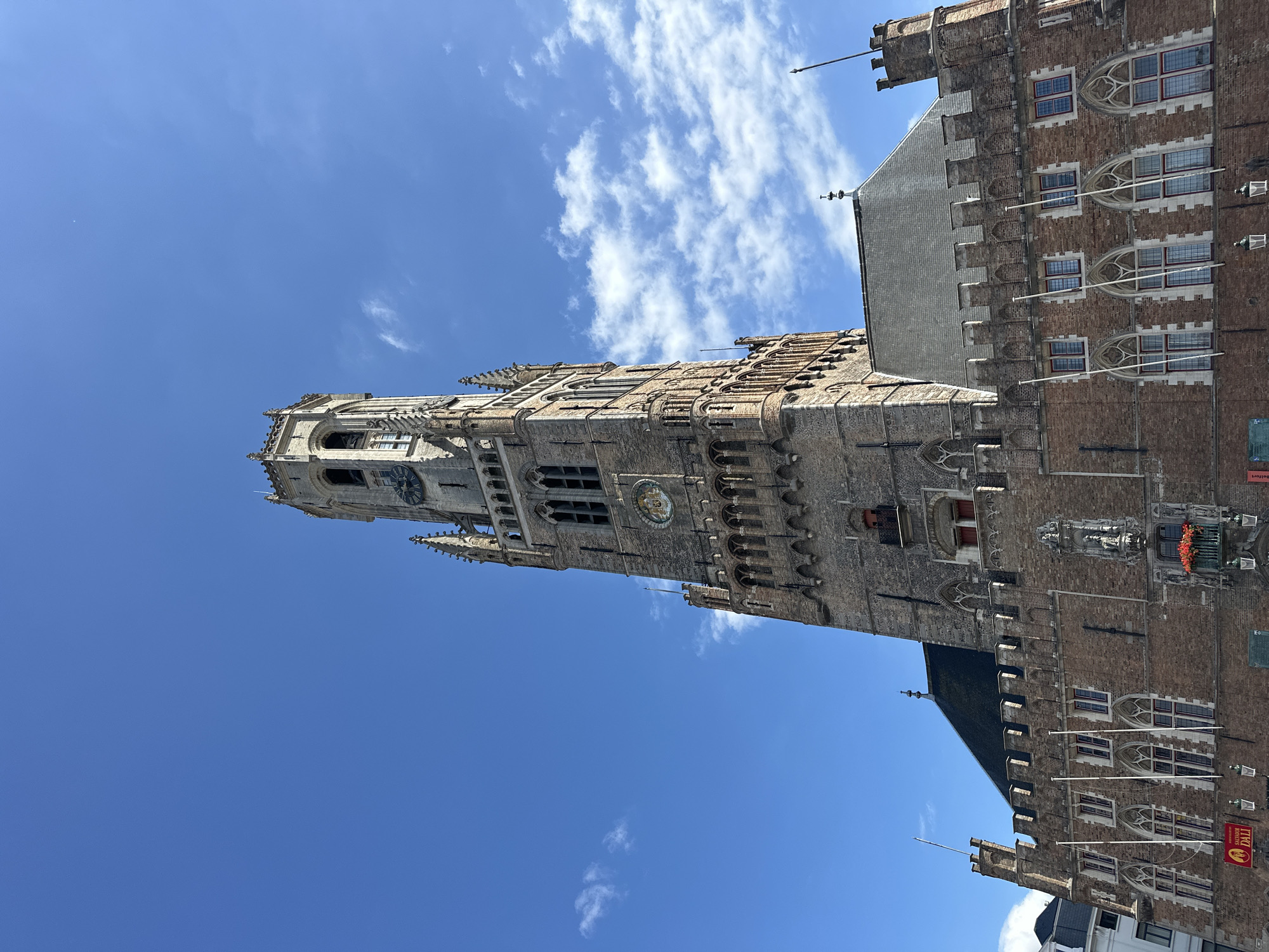 The image captures a low-angle view of the Belfry of Bruges, a medieval bell tower located in the Market Square (Markt) of Bruges, Belgium. The tower is constructed of brick and stone, with a pointed spire at its peak. It features numerous arched windows, decorative stonework, and a large clock face visible on its upper section. Below the tower, a long, multi-story building with similar architectural elements, including arched windows and crenellated parapets, stretches across the frame. The sky above is a clear blue with scattered white clouds. A portion of a red banner with white text and a Salvador Dalí advertisement is visible at the lower left. The scene appears to be a sunny day.