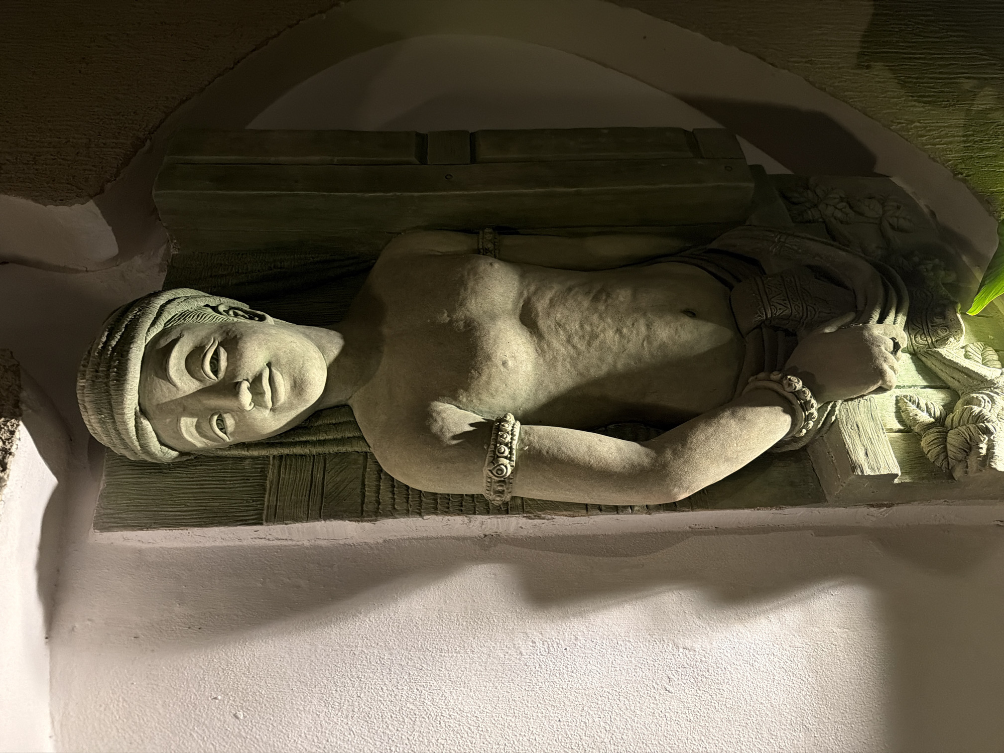 This image, taken in Ao Phai, Thailand, presents a close-up of a stone relief sculpture depicting a smiling, bare-chested female figure integrated into a textured wall. The sculpture portrays the figure with a wrapped head covering, narrow eyes, a small nose, and full lips. Her left arm, bent at the elbow, wears a patterned circular armlet. The torso displays subtle anatomical details and textured skin, while the lower body is draped in a patterned, wrapped garment. Behind the figure, the relief includes a textured panel with vertical striations. The scene is illuminated by artificial light from the lower left, casting prominent shadows on the surrounding light-colored, textured wall to the left and highlighting the sculpture's features. A darker, roughly textured, and curving wall is visible to the right of the sculpture. A faint green light emanates from the bottom edge of the frame, possibly from nearby foliage or a colored light source.