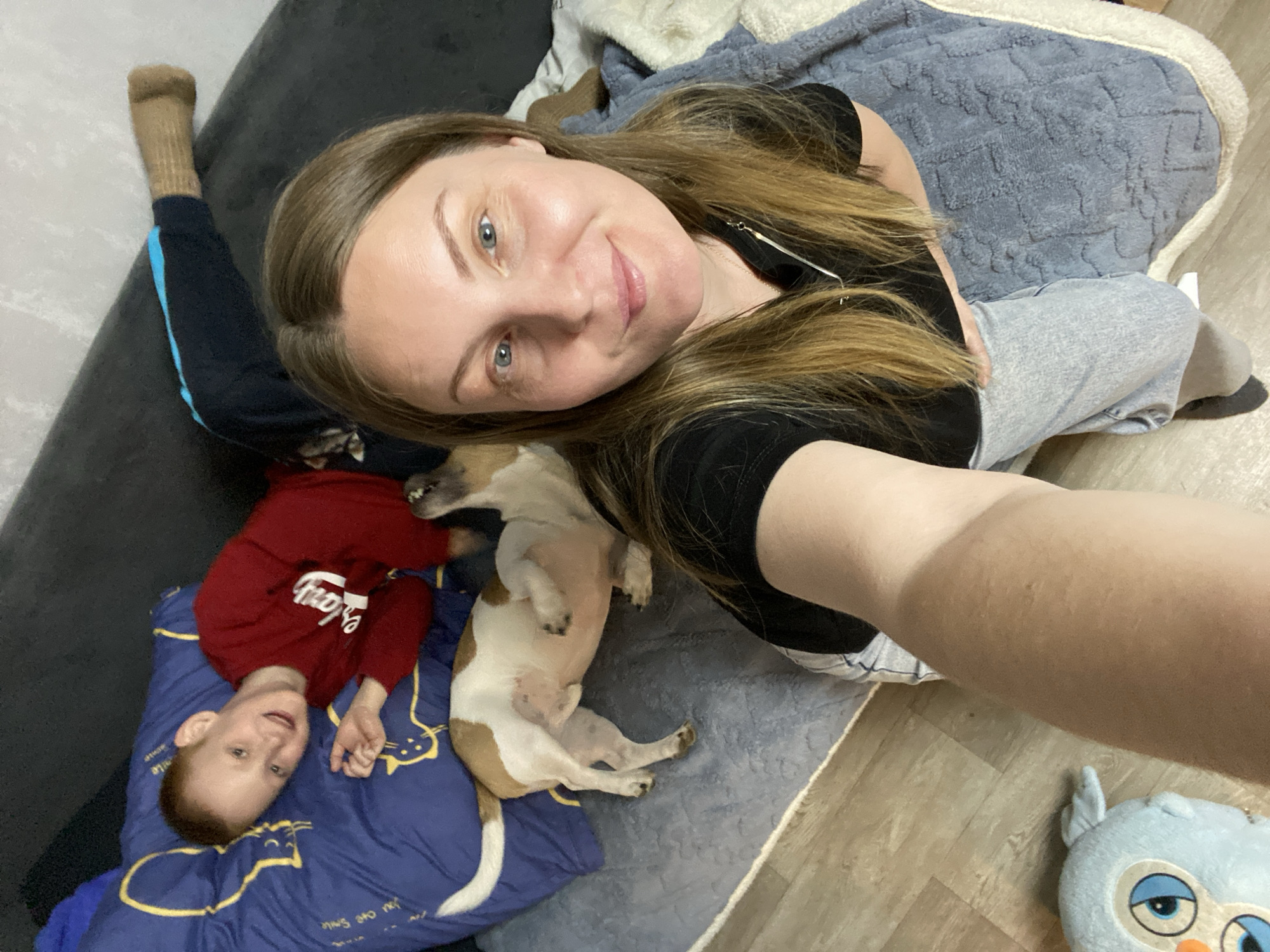 A woman is taking a selfie from a top-down perspective. She is lying on a grey blanket or cushion on the floor. She is wearing a black t-shirt and light-colored trousers. Next to her, there is a white and brown dog, lying on its back. A young boy, dressed in a red sweatshirt with white text, is lying on a blue cushion with yellow cat designs. To the right of the woman, there is a blue stuffed owl toy. The floor is a light wooden color. A small portion of a couch can be seen in the top left of the frame. The location and city cannot be confirmed from the image.