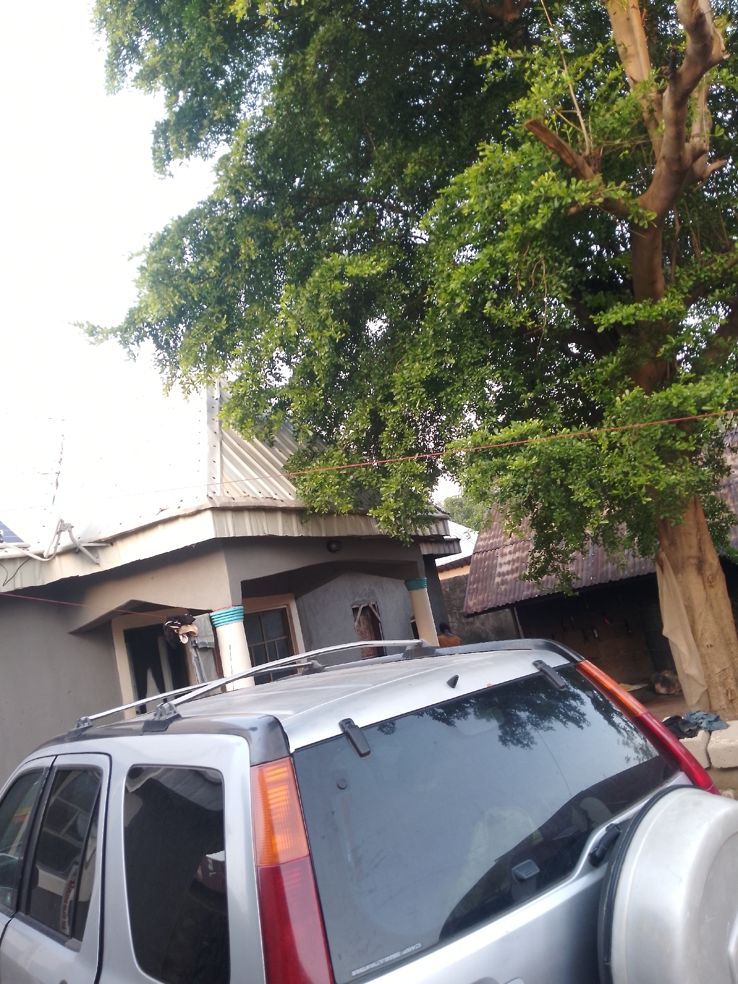 A silver SUV is parked in front of a building with a corrugated metal roof. A large tree with lush green leaves overhangs the scene, casting shadows. The building appears to be residential, with windows and a covered entrance. The sky is bright and overcast.
