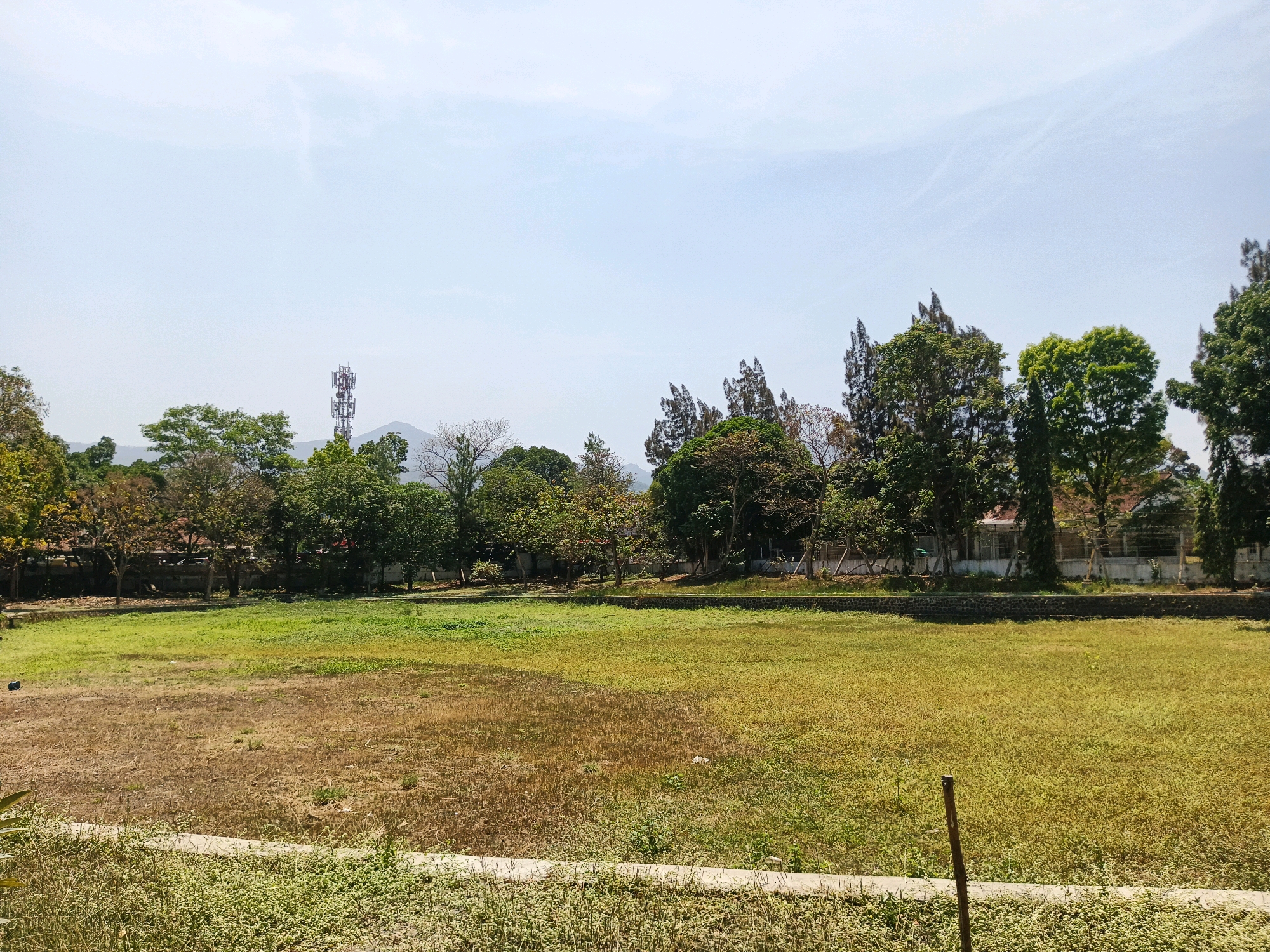 The image shows an outdoor scene with a large, open field. The field is a mix of dry, brown grass in the foreground and greener grass in the mid-ground. A concrete path runs along the bottom edge of the image, bordered by low-lying vegetation. In the background, a line of trees with various types of foliage separates the field from buildings. Beyond the buildings, a cell tower is visible against a pale sky with some wispy clouds. In the far distance, hills or mountains can be seen. The lighting suggests it is daytime, likely late morning or early afternoon. There are no people or animals visible in the scene. The environment appears to be a park, sports field, or undeveloped land within a semi-urban or suburban area, potentially on the outskirts of Bandung City, Indonesia, given the presence of the cell tower and distant hills typical of the region.