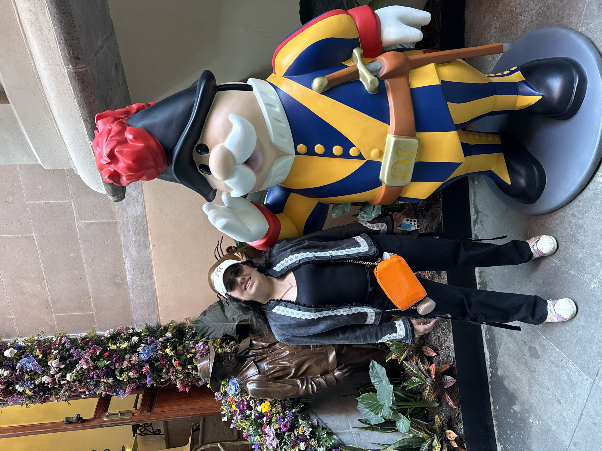 A woman stands next to a large statue of a man in uniform, with a smaller bronze statue in the background amidst flowers and plants. The woman is wearing sunglasses, a white cap, a black top, a black jacket with white lace trim, black pants, and white shoes. She has an orange purse on a chain across her body. The large statue is a cartoonish figure in a blue and yellow uniform with a black hat and a red pompom. The setting appears to be an interior space with stone walls and flooring, possibly in Mexico City, Mexico. There are flowers and plants along the wall and around the bronze statue.