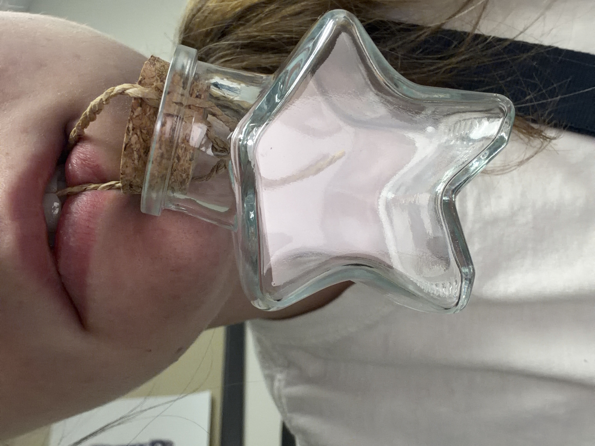 A close-up, waist-up shot shows a person's mouth biting a woven twine handle attached to a star-shaped glass bottle filled with a light pink substance. The bottle has a cork stopper. The person is wearing a white t-shirt. The background is blurred and shows a wall and some shelving. The image was taken in Beaverton, Oregon, United States of America.
