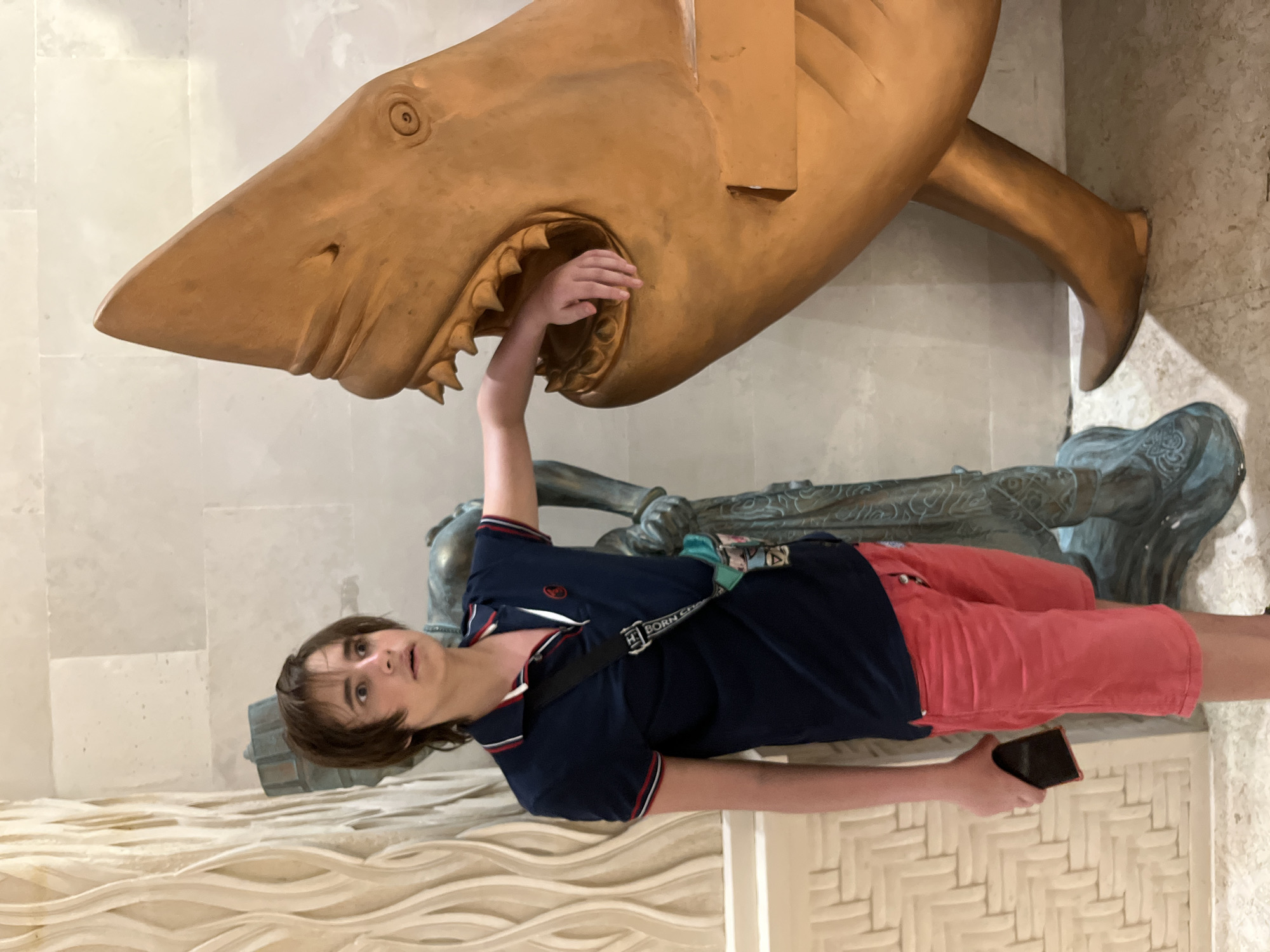 A young person stands in front of a large, golden shark sculpture with its mouth open. The person, wearing a dark blue polo shirt with red and white trim and red shorts, holds a dark-colored smartphone in their left hand. Their right arm is extended towards the shark's open mouth, with their hand inside. Behind the person, a bronze statue of a person is visible, partially obscured by the shark sculpture. The background consists of a light-colored textured wall and tiled floor. This scene appears to be captured at the Bali Art Market (Pasar Seni Bali) in Sukawati, Indonesia, known for its vibrant collection of sculptures and artwork.