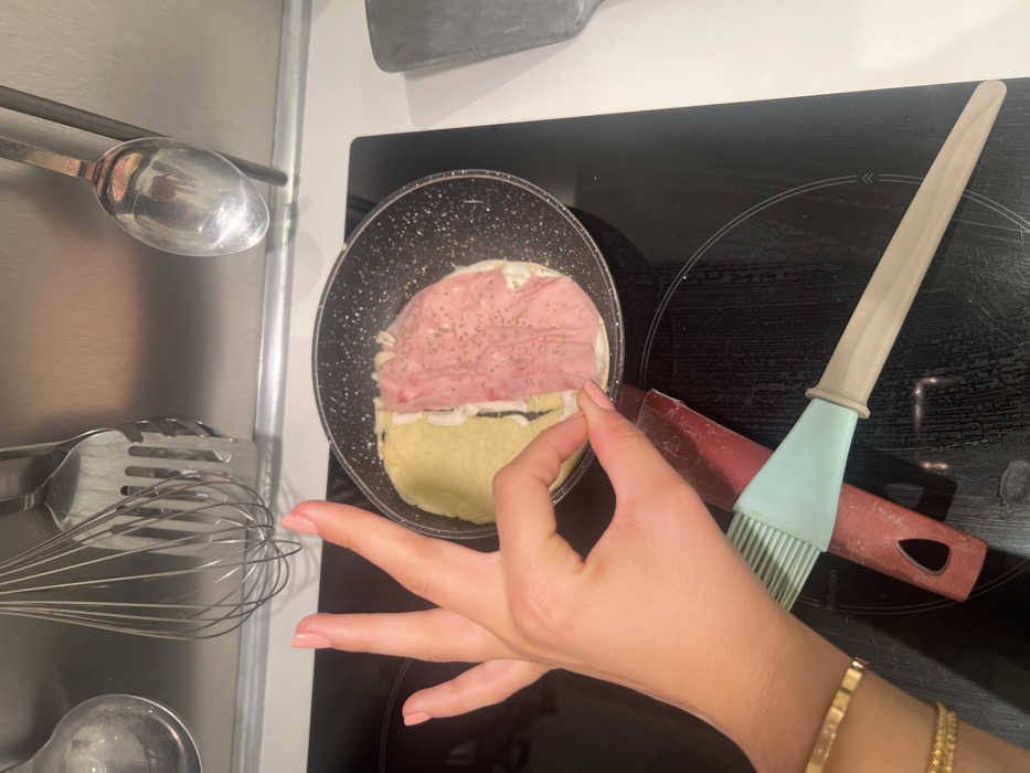 A close-up shot shows a frying pan on a black stovetop, cooking what appears to be a crepe or pancake with ham and a white sauce. A cooking brush lies next to the pan. A hand with manicured nails reaches towards the food. Utensils hang on a rack on the left side of the frame. A gold bracelet is visible on the wrist of the person whose hand is reaching into the frame.