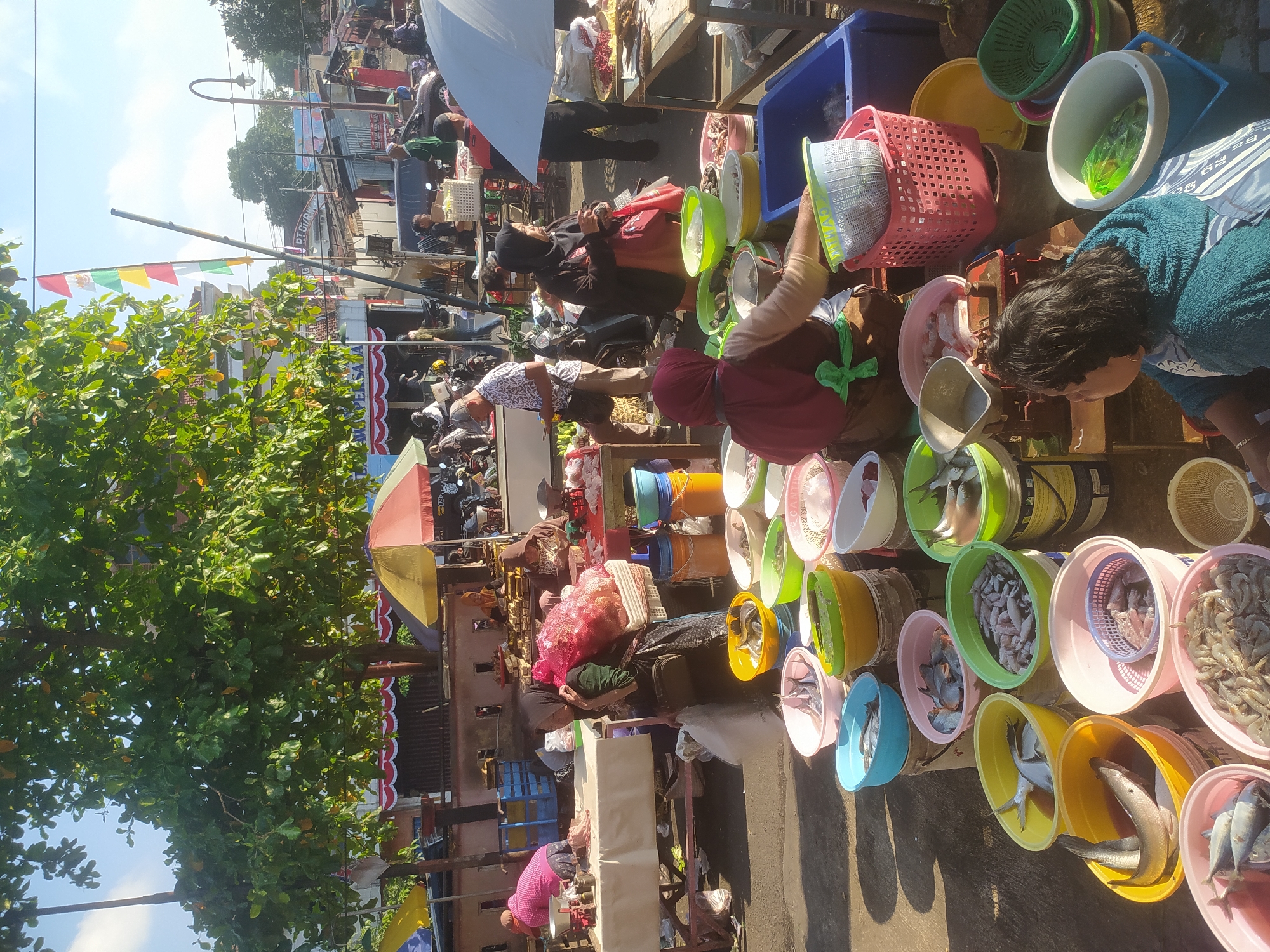 This is a street market scene in Kupang, Indonesia. The foreground is dominated by a variety of colorful plastic basins and buckets, many filled with fresh fish and shrimp. Several women are tending to the stalls. One woman in the center foreground, wearing a teal hooded garment, is looking towards the right. To her left, a woman in a red hijab is sorting through a bin of shrimp. Another woman, further back, wearing a black headscarf and pink shirt, is weighing fish on a scale. Behind the stalls, there are more market activities with people milling about, some carrying goods. Various stalls, umbrellas, and temporary structures suggest a busy marketplace. The overall environment appears to be an outdoor urban setting, with evidence of trees and buildings in the background. The bright sunlight indicates it is daytime.