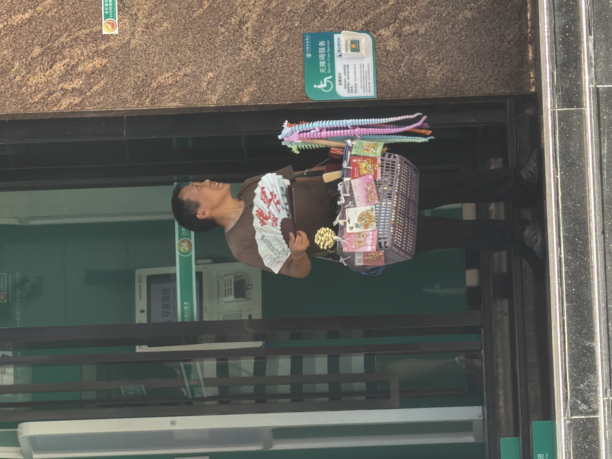 The image depicts a man standing in front of a building entrance in the Yuzhong District, China. He is wearing a brown t-shirt and black pants, holding a folded fan in his left hand and a basket filled with small toys and trinkets in his right. The basket contains items such as colorful segmented toys, small packages with Chinese writing, and what appear to be charms or ornaments. The man is looking to his right, with a slight smile. Behind him, a glass door leads into a room with green walls and an ATM machine visible, displaying Chinese text. A sign with a wheelchair symbol and Chinese text indicating "Barrier-Free Service" is affixed to the brown wall on the right side of the image. The man is standing on a set of dark steps leading up to the building entrance.