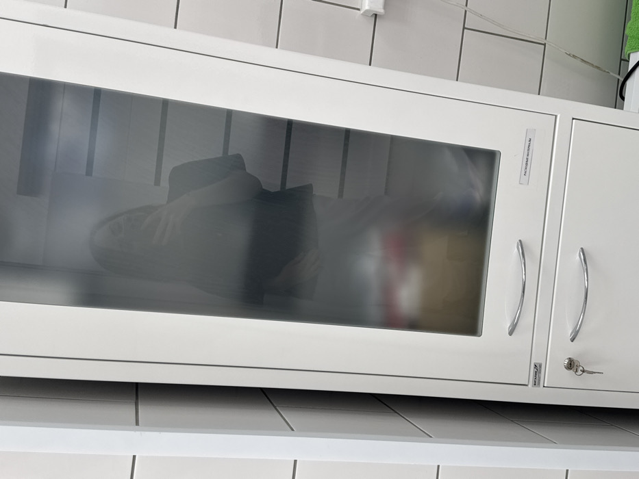 A white cabinet with a transparent glass door dominates the frame. A second, opaque white cabinet sits adjacent. The cabinet doors have curved silver handles and a visible keylock. An object, possibly clothing or fabric, is visible inside the glass-fronted cabinet. The cabinet sits against a tiled wall. A thin white shelf is visible below the cabinet. A small white electrical outlet is visible near the top of the frame.