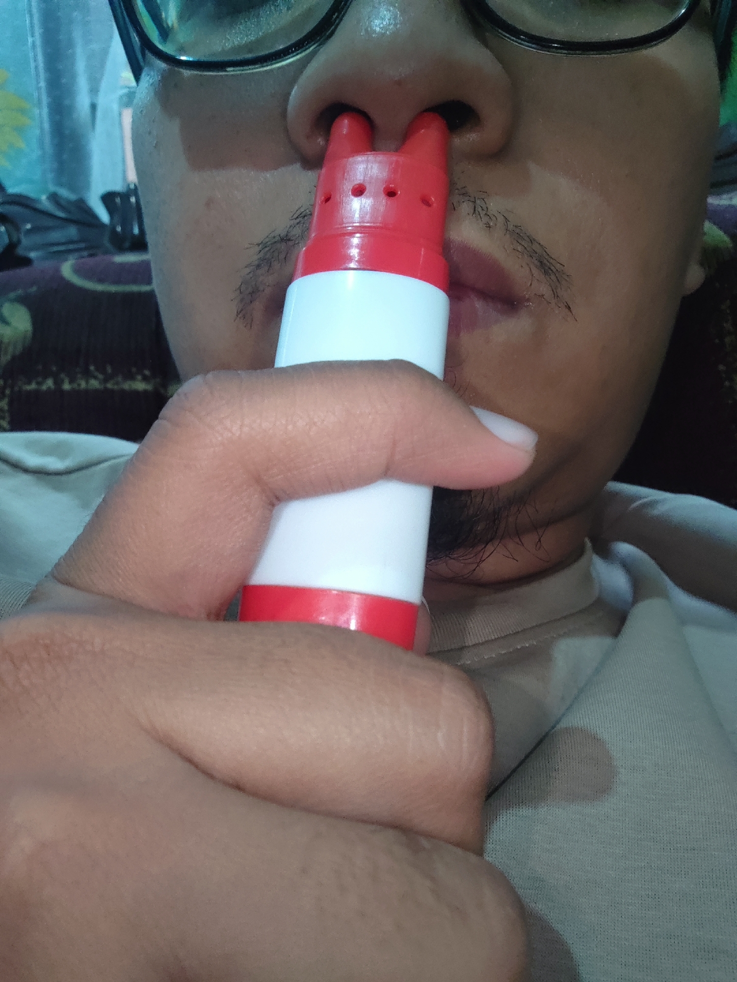 A close-up, head-and-shoulders shot shows a person's face and hand in the city of Bondowoso, Indonesia. The person is wearing glasses and has a light mustache and beard. They are holding a red and white nasal spray device to their nose with their right hand. The device has several small holes on the red tip. The person is wearing a light-colored t-shirt. The background is slightly blurred and appears to be indoors, with a patterned fabric on a couch visible. The lighting suggests an indoor setting, possibly in a home environment.