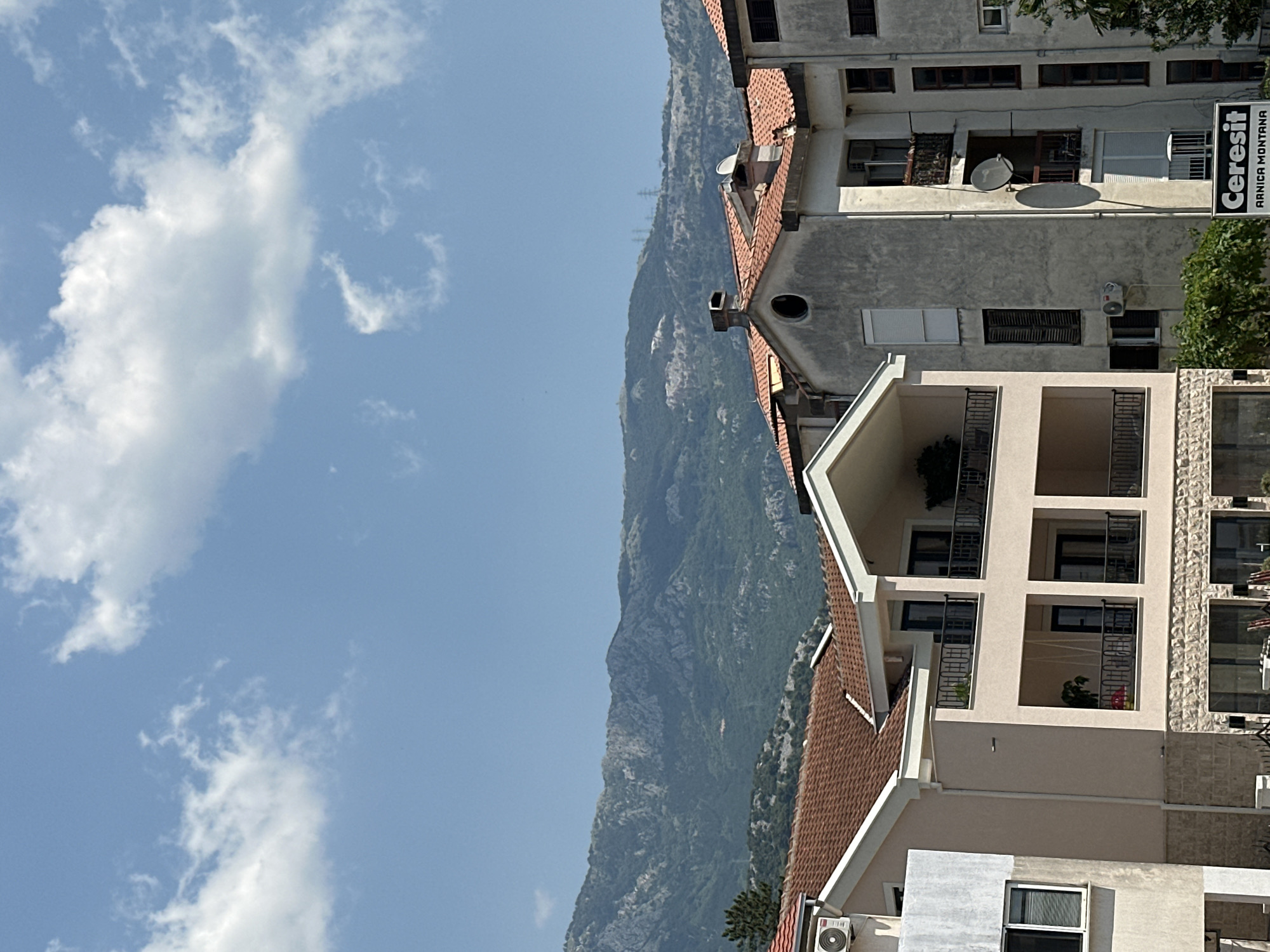 This is a vertical, outdoor, medium shot of a mountainous landscape with buildings in the foreground and a cloudy sky in the background. The scene is set in Budva, Montenegro, on a clear, sunny day.
The dominant subject is a vast, rugged mountain range covered in dense green vegetation, rising steeply into the sky. In the foreground, several multi-story buildings are visible. These buildings are architecturally varied, with some featuring red-tiled roofs and others with modern facades. Balconies, windows, and air conditioning units are noticeable architectural details. One building has a prominent sign that reads "Ceresit ARNICA MONTANA". The sky is a bright blue, populated by scattered white clouds.
No people or specific activities are depicted. The overall impression is one of a scenic natural environment juxtaposed with urban development. The lighting suggests it is daytime, likely late morning or early afternoon.