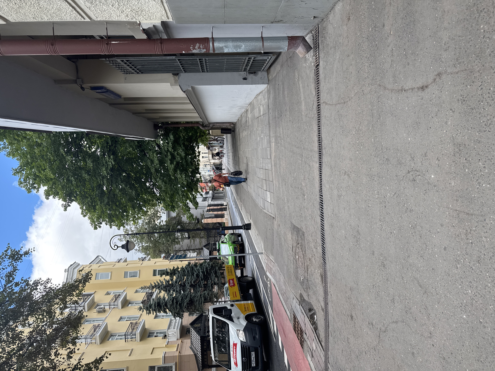 The image depicts a street scene in Moscow, Russia, on a cloudy day. In the foreground, a gray asphalt sidewalk is visible, with metal grates for drainage running along its edge. To the left, a street with a red and white striped lane, likely for bicycles or pedestrian crossing, is seen. A white utility van with a yellow trailer is parked on the street, displaying Russian license plates. A green car is also present on the street.
Further down the sidewalk, a man is walking away from the camera, carrying two bags. Behind him, other pedestrians are visible, as well as lampposts and trees lining the street. The buildings flanking the street are multi-story structures with yellow and gray facades, featuring balconies and windows. The architecture suggests an urban environment. The overall impression is of a typical day in a residential or commercial district of Moscow.