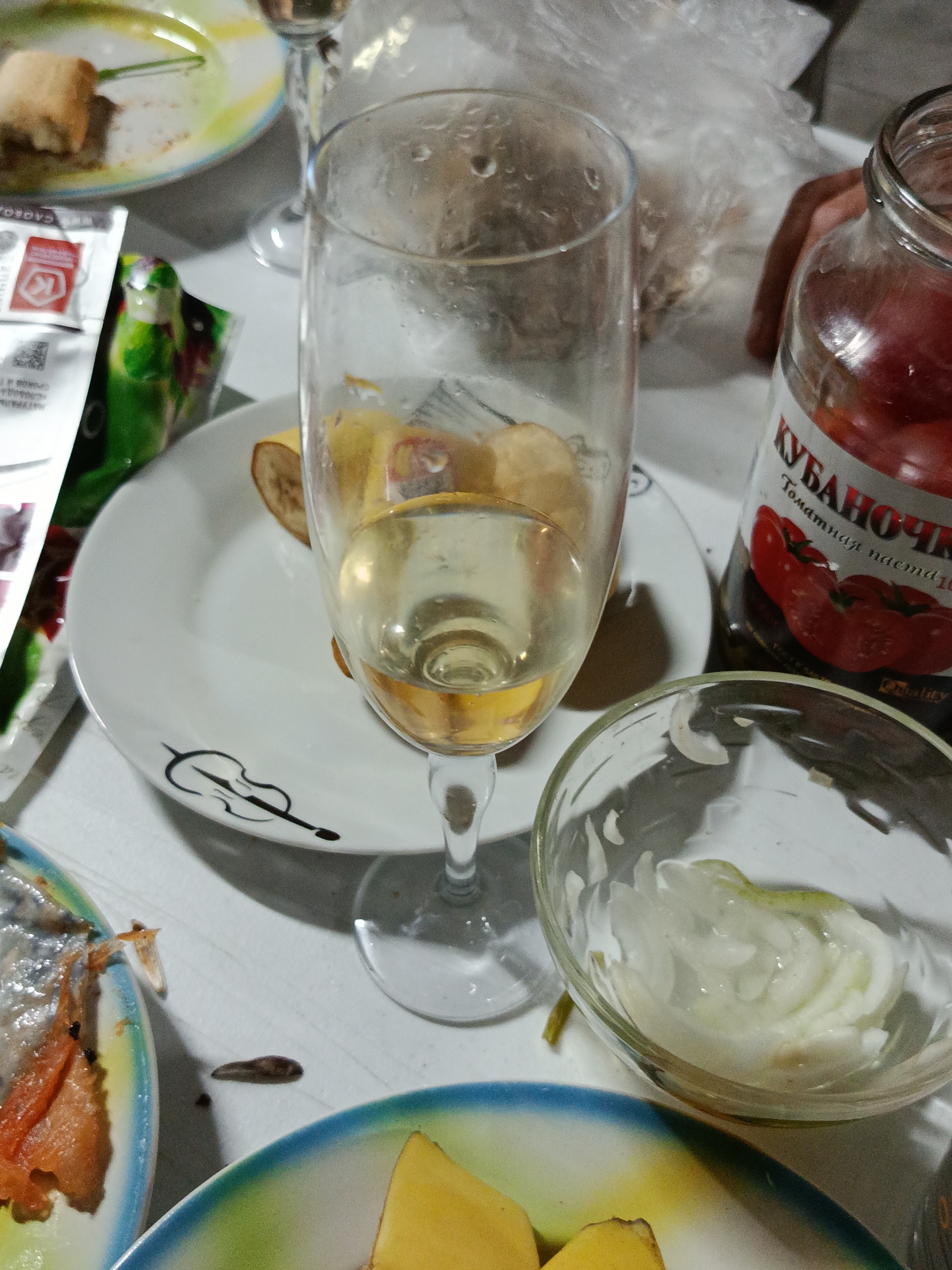 The image depicts a table setting in Novotitarovskaya, Russia, with various food items and drinks. In the foreground, a partially eaten plate of what appears to be fish or seafood is visible. Next to it, a small, dark, elongated object lies on the table, possibly a piece of debris or food. A glass bowl contains sliced onions. Further back, a white plate with a violin design holds banana slices. A wine glass filled with a pale yellow liquid stands prominently. To the right, a glass jar labeled "КУБАНОЧКА" (Kubanochka), containing red tomatoes, is partially obscured. The background shows plastic packaging and a person's hand. The overall scene suggests a casual meal or gathering.