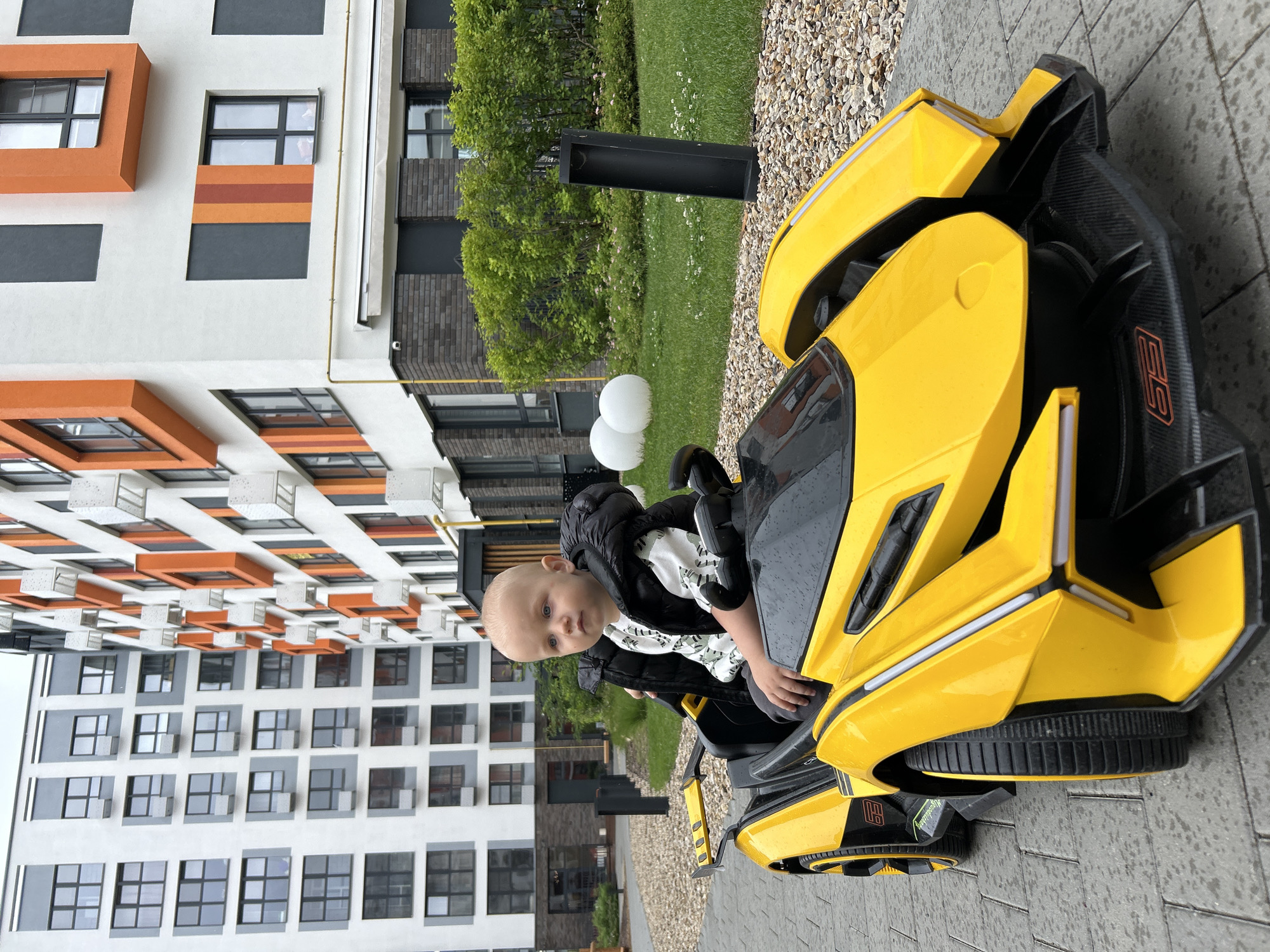 A fair-skinned toddler with blue eyes is sitting in a bright yellow toy electric car with black accents and black tires. The child is wearing a black puffer vest over a white t-shirt with a camouflage print. The car is parked on a paved walkway bordered by small, light-colored stones. In the background, there is a modern apartment building with white, gray, and orange facades and numerous windows. Green grass and dense bushes are visible in front of the building, along with two large, white spherical objects and a tall, black rectangular planter. The overall scene appears to be an outdoor residential area.