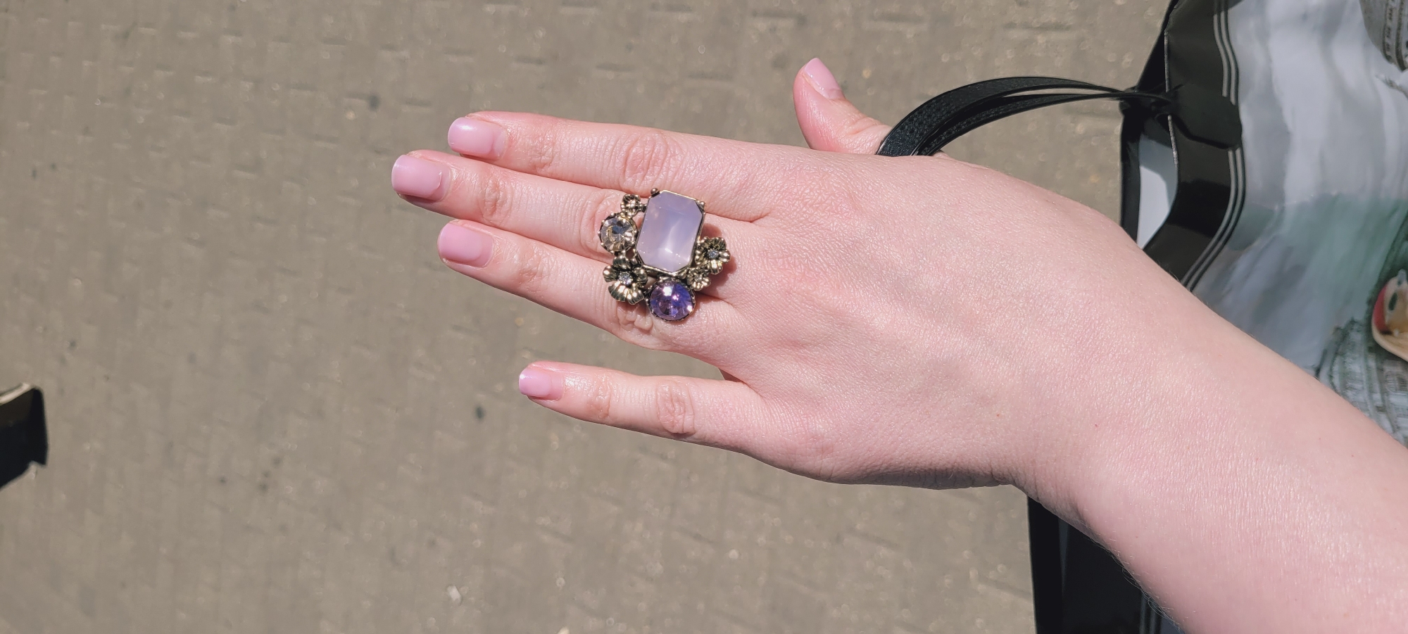 A human left hand is depicted, viewed from above, holding the black handle of a glossy plastic shopping bag. The hand exhibits light-toned skin, with all visible fingernails painted in a light pink polish. An ornate ring is worn on the middle finger. The ring features a central, rectangular, pale purple stone, bordered by a smaller, round, deep purple stone on the left, and several smaller, clear faceted stones set within an antique gold or bronze-toned metalwork resembling floral or leaf motifs. The partially visible shopping bag displays a monochrome print of architectural structures, including domed buildings, with a small object wearing a red cap visible at the bottom of the bag's print. The background consists of a light grey-brown paved surface, possibly cobblestones or bricks, indicating an outdoor setting under direct sunlight. A blurred dark object is present in the upper background. The scene is located in Unknown, Unknown.
