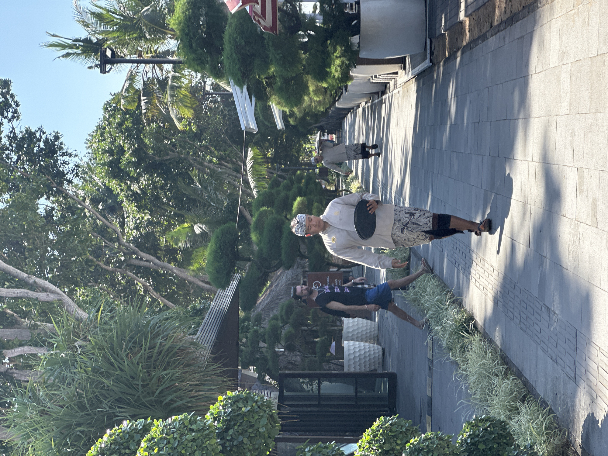In Denpasar, Indonesia, a person in traditional Balinese attire, including a headcloth and patterned sarong, walks along a paved walkway lined with manicured trees in planters. The individual carries a dark, round tray. To the left, another person wearing a black t-shirt and blue shorts walks away from the camera. In the background, more trees and buildings are visible under a clear blue sky. The sun casts shadows on the walkway, indicating a bright day.