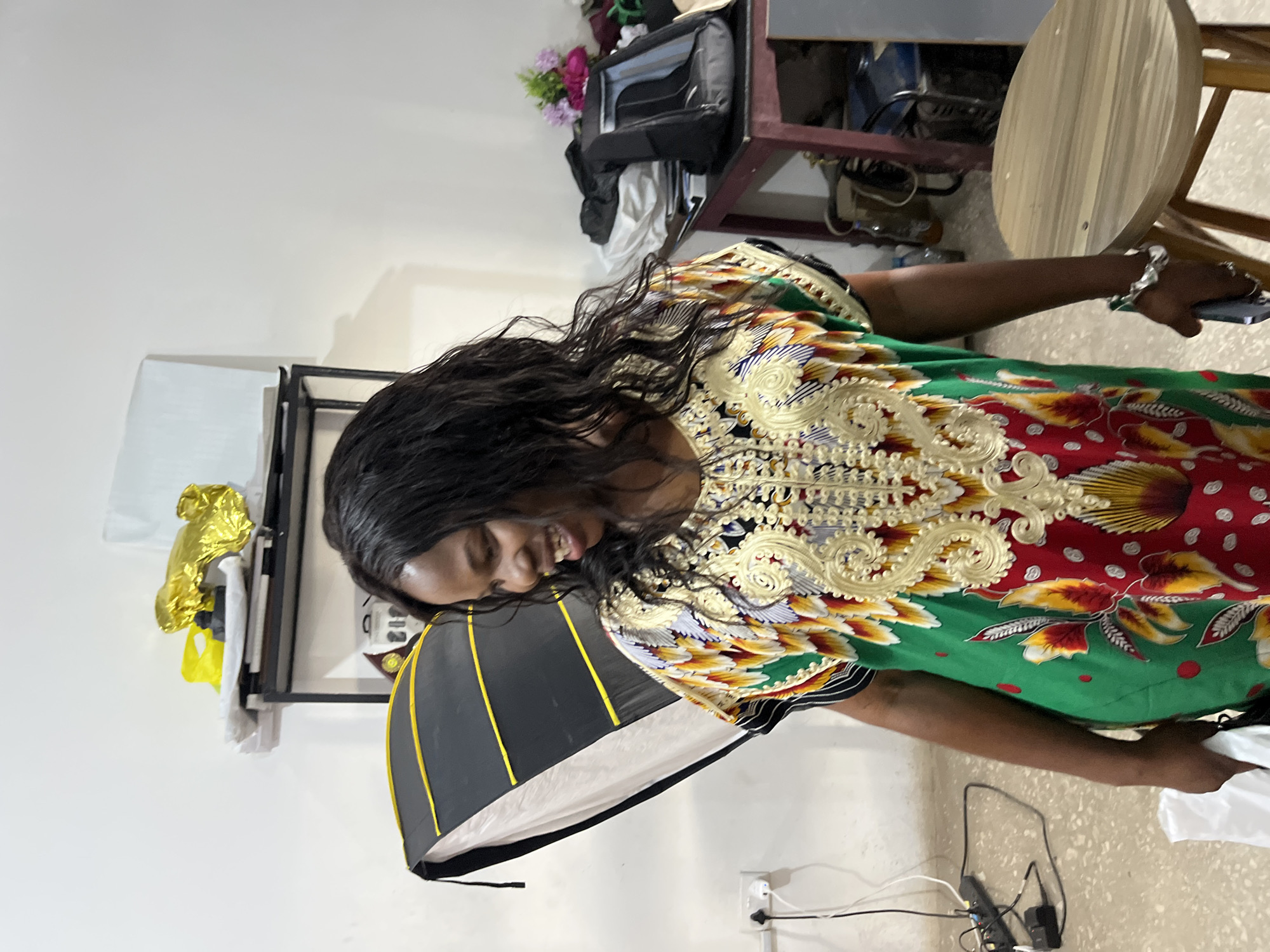 A Black woman with long, dark, wavy hair, wearing a brightly colored dress with a green base and intricate gold, red, yellow, and white floral patterns, is standing and laughing. She is looking to her right. She is wearing a silver bracelet and several silver rings. In her right hand, she is holding a dark-colored smartphone. In her left hand, she is holding a white bag.
The background shows a room that appears to be a studio or storage space. To the left, a large, collapsible photography softbox is leaning against a wall. A power strip with multiple plugs is attached to the wall near the floor. Further back, a black metal shelf unit holds various items, including a crumpled gold object, a yellow bag, and white materials. To the right, a dark wooden table with a reddish hue holds a black backpack, some folded clothing, and a small bouquet of pink and purple flowers. A cylindrical wooden stool with light-colored wood grain is positioned in the foreground to the right. The walls are white, and the floor appears to be made of speckled concrete.
The image was taken inside a room in Gura Top, Nigeria.