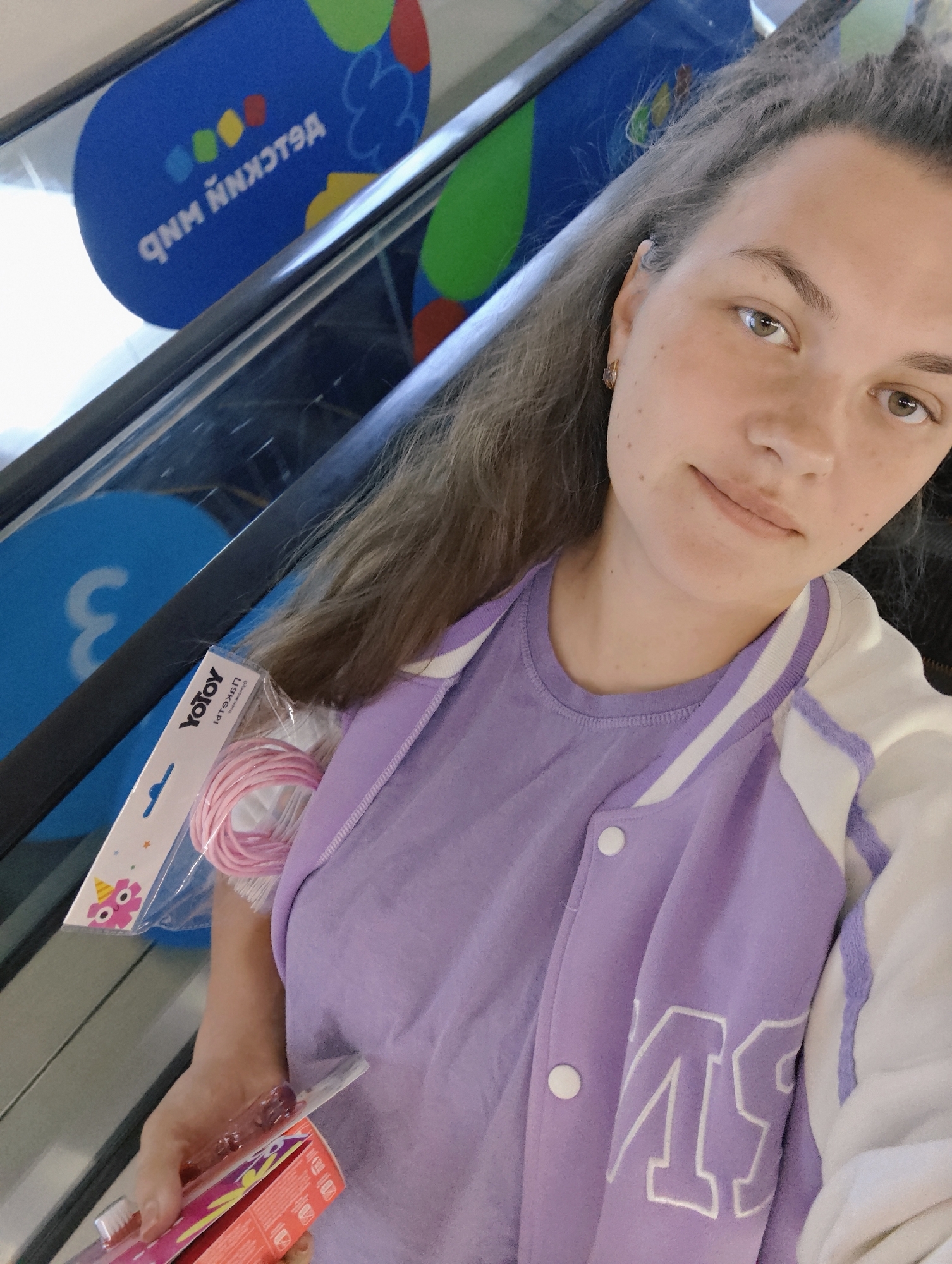 A woman is taking a selfie while riding an escalator. She is holding a package labeled "YoToy" and a toothbrush. She wears a light purple T-shirt and a jacket with white sleeves and light purple accents. The escalator has signage with the Cyrillic text "Я люблю Чайковский", which translates to "I love Chaikovsky," suggesting the location is in the city of Chaikovsky, Russia.