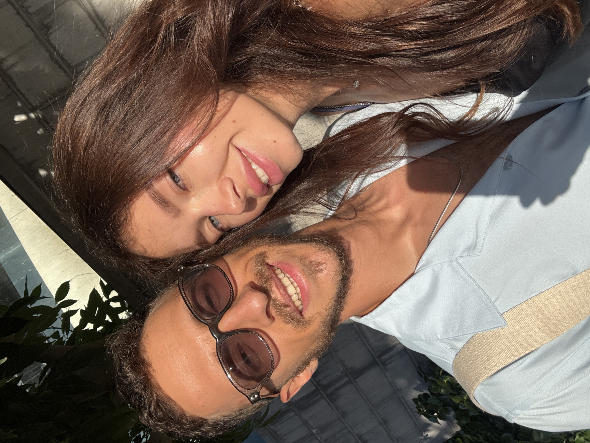 A man and woman are posing for a selfie outdoors in Tel Aviv, Israel. The man, on the left, wears sunglasses and a light blue collared shirt. He has a beard and short, dark hair. The woman, on the right, has long, dark brown hair and is smiling. They are both looking at the camera. The background appears to be an urban setting with a brick wall and foliage. The lighting suggests it is daytime, possibly late afternoon, with sunlight casting shadows.