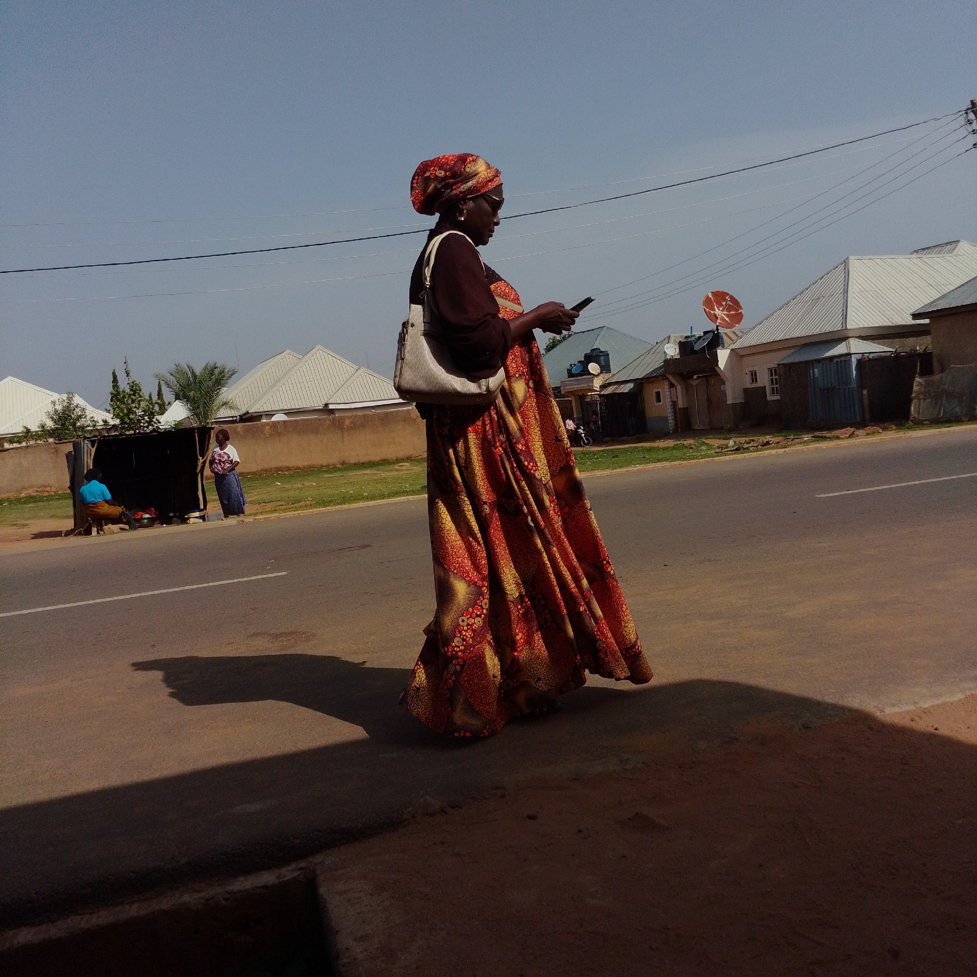 A woman in a colorful, flowing dress and headscarf walks along the side of a paved road in Gombe, Nigeria. She is holding a mobile phone in her right hand, and a light-colored shoulder bag is slung across her left shoulder. The woman is in profile, facing right, and appears to be looking at her phone. Behind her, on the opposite side of the road, are several buildings with corrugated metal roofs, some with satellite dishes. There is also a small open-air stall with two individuals visible. The environment appears to be urban or semi-urban, with power lines visible overhead. The road is asphalt with a white line marking the lane. The lighting suggests it is daytime, with shadows cast on the road.