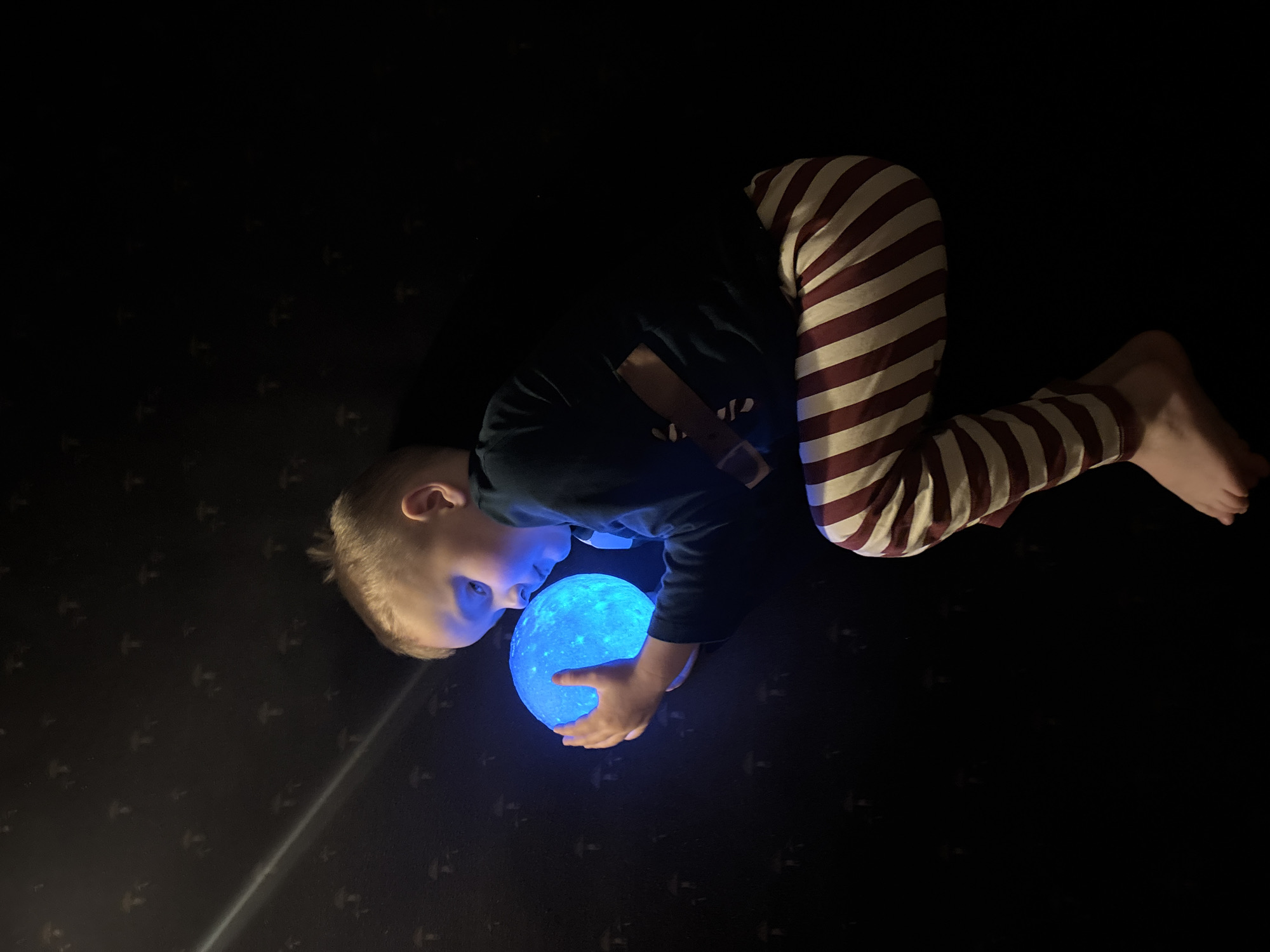 A young child is lying on a dark surface with a patterned floor. The child is wearing striped pajama pants and a dark long-sleeved shirt. They are holding a glowing blue orb in their hands and looking at it intently. It's impossible to determine the location in Chaikovsky, Russia based on the provided image.
