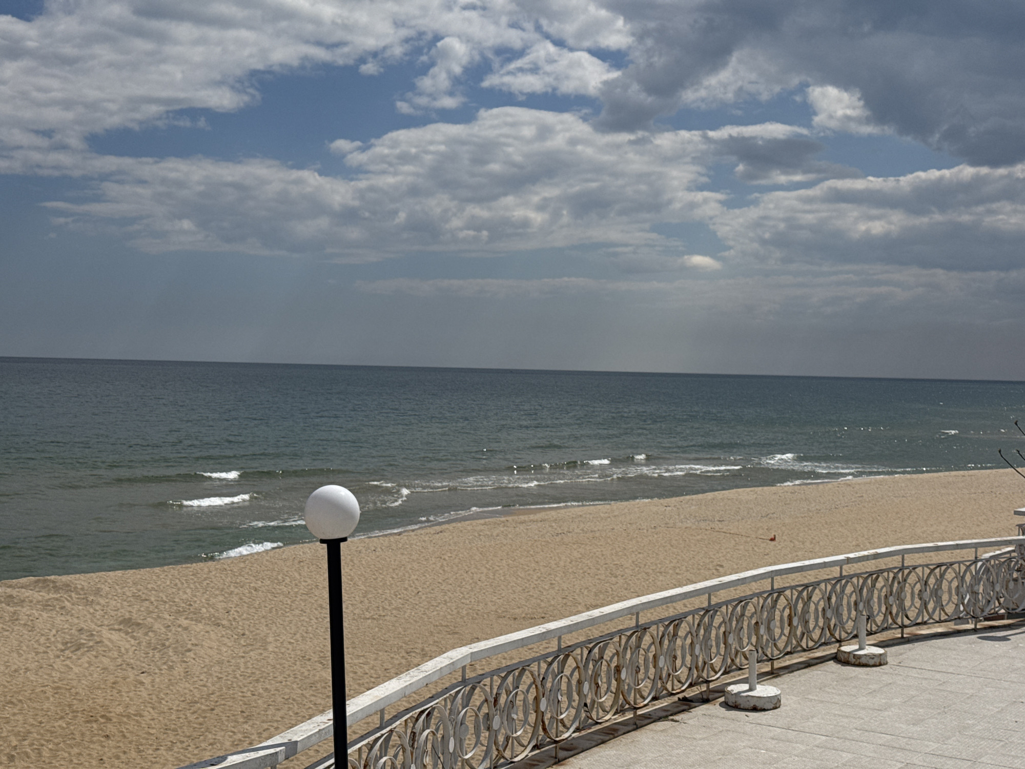 The image shows a beach scene in Varna, Bulgaria. The view is from an elevated platform with a decorative white railing. A white spherical lamp post stands near the railing. The beach is wide and sandy, with gentle waves lapping the shore. The sea is a muted blue-grey color. The sky is overcast with varied cloud formations.