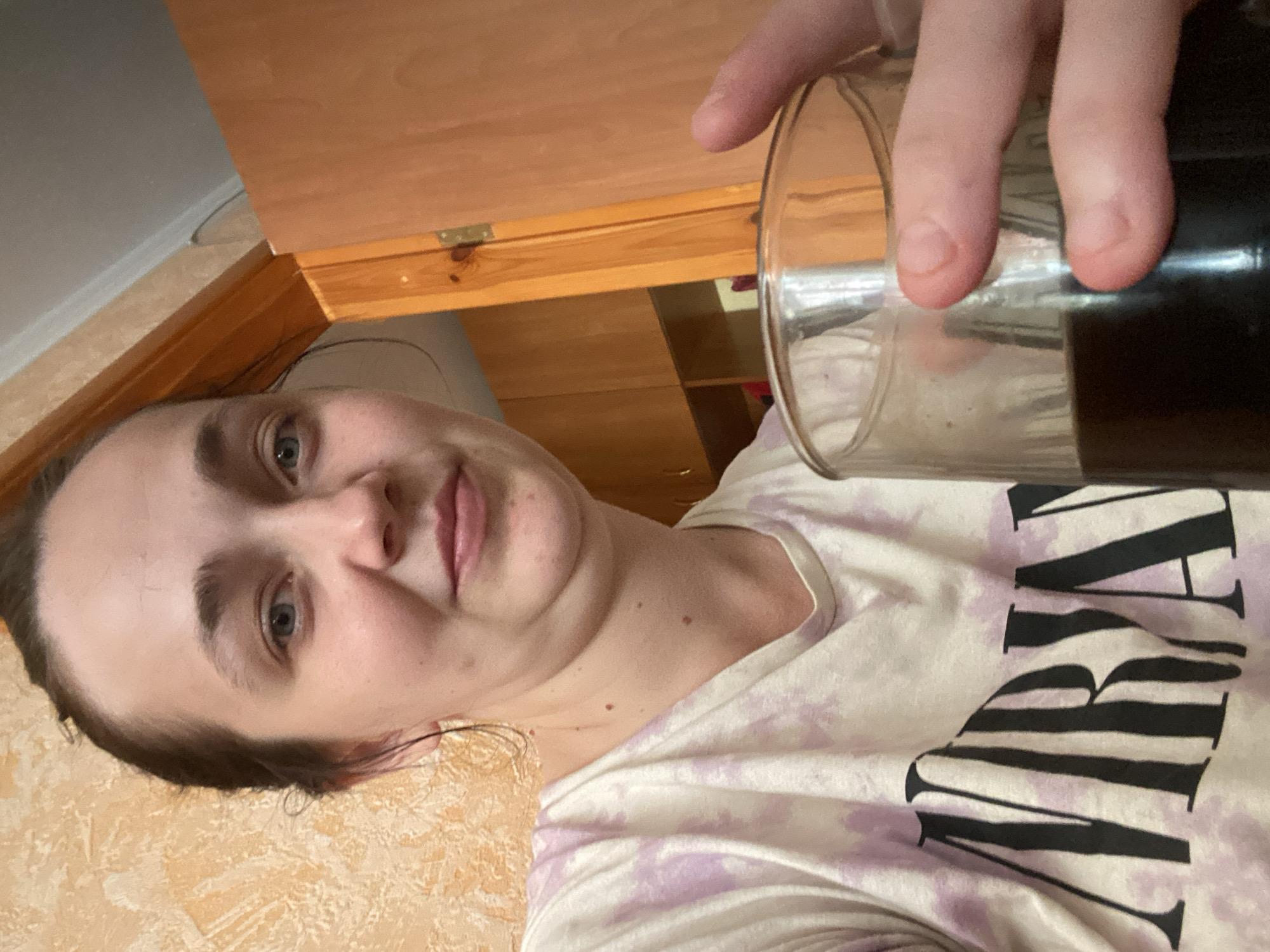 A woman is holding a glass of dark liquid in her right hand. She is wearing a tie-dye t-shirt with the word "NIRVANA" in black block letters across the front. The background appears to be an indoor setting with textured orange wallpaper and wooden cabinetry. The woman's hair is dark brown and pulled back. Her eyes are blue, and she is looking directly at the camera with a slight smile.