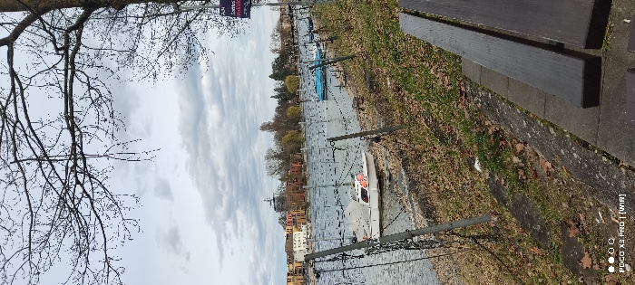 This static, vertical image depicts a tranquil waterfront scene in Constance, Germany, under a heavily overcast sky. The immediate foreground on the right features a dark-brown wooden slatted bench situated on a concrete path. Adjacent to this path is a gently sloping grassy embankment, sparsely covered with green blades and numerous scattered dry brown leaves. A barrier, constructed from grey metal posts connected by taut wire strands, separates the embankment from the water.
In the mid-ground, a calm body of water, consistent with Lake Constance or a connected waterway, stretches horizontally across the frame. Numerous slender, dark vertical poles are distributed throughout the water, likely serving as mooring points. Two distinct recreational boats are visible: a white cabin cruiser with a grey top and an orange detail near the stern is moored on the left, and a smaller, bright blue open boat is moored further to the right.
The background reveals the distant city shoreline of Constance. It features a cluster of multi-story buildings with red-tiled roofs and facades in shades of white, beige, and terracotta. Prominently positioned behind these structures, slightly left of center, is a building distinguished by a dark, possibly cylindrical or conical tower. The far bank is lined with leafless deciduous trees. The upper portion of the frame is filled with the intricate, bare branches of a large tree, silhouetted against a uniform expanse of grey and white clouds. No individuals are present, and no activity is observed in the scene.