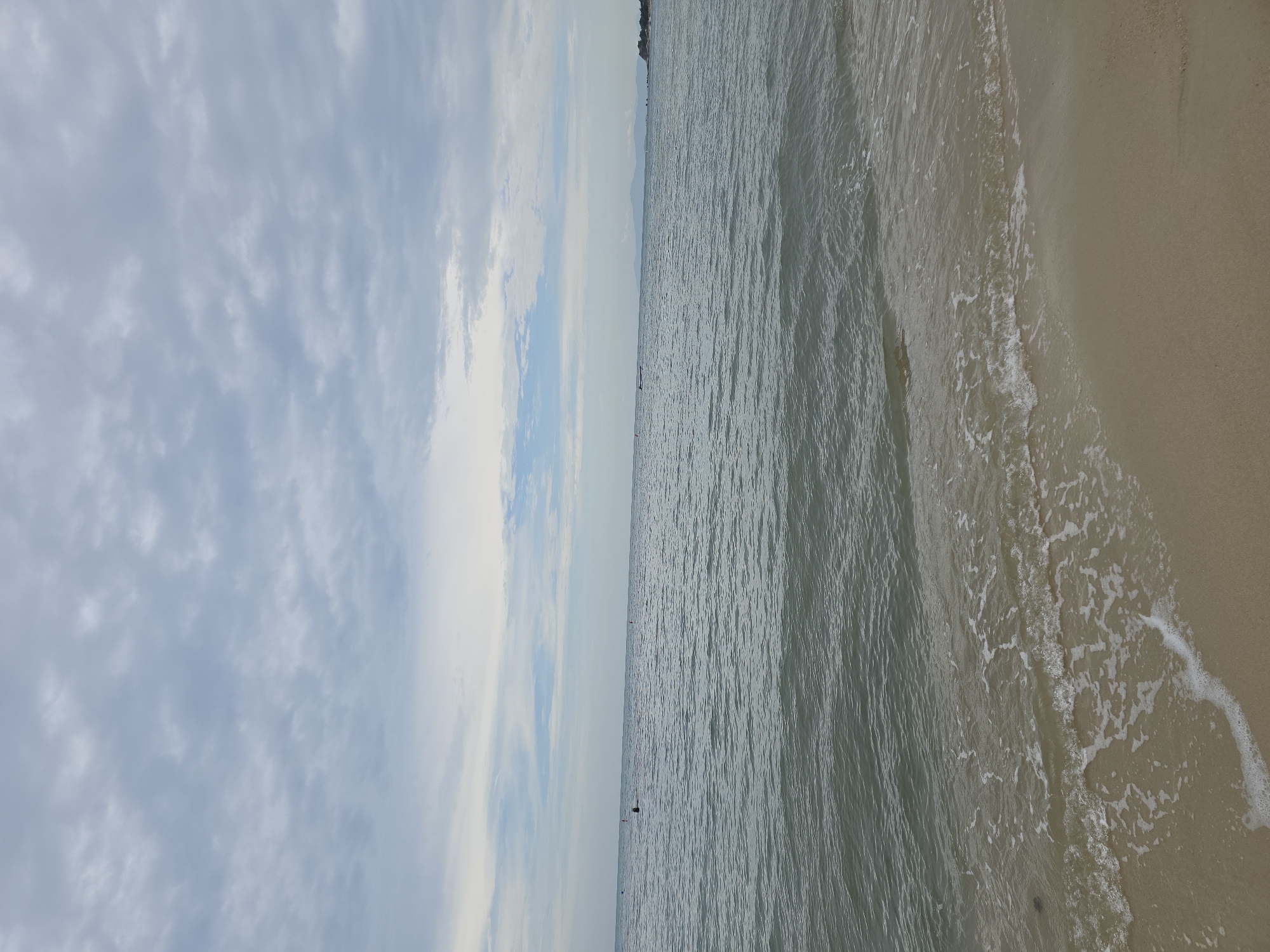 The image depicts a serene beach scene with a calm sea under a cloudy sky. The sand in the foreground is a light tan color, and a gentle wave is lapping at the shore, creating a frothy white edge. The sea stretches out to the horizon, with subtle ripples on its surface. In the distance, faint landmasses can be seen along the coastline, dotted with some structures. Several small boats or buoys are visible on the water, and a few figures are swimming or wading in the sea. The sky is filled with a pattern of gray and white clouds, suggesting an overcast day. The overall impression is one of tranquility and natural beauty.