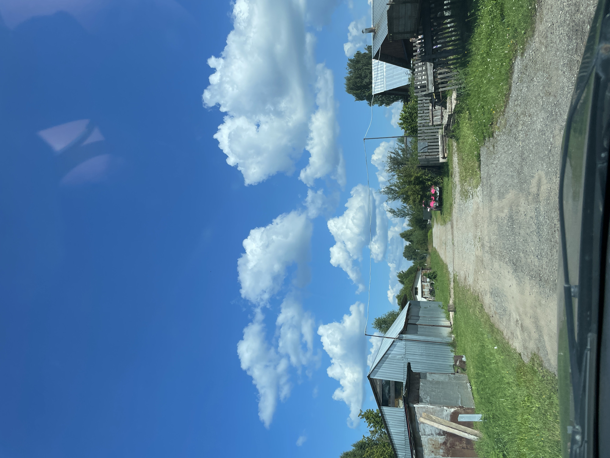 The image shows a clear blue sky with scattered white clouds above a dirt road in the village of Дресвяново, Russia. The road is flanked by grass and vegetation. On the left side of the road, there is a corrugated metal building, possibly a garage or storage facility, next to a smaller structure. On the right side, there are wooden fences, some old and weathered, and a few residential buildings with metal roofs. Further down the road, a pink vehicle is parked, and more trees and vegetation are visible. The scene appears to be an outdoor, rural environment on a sunny day.