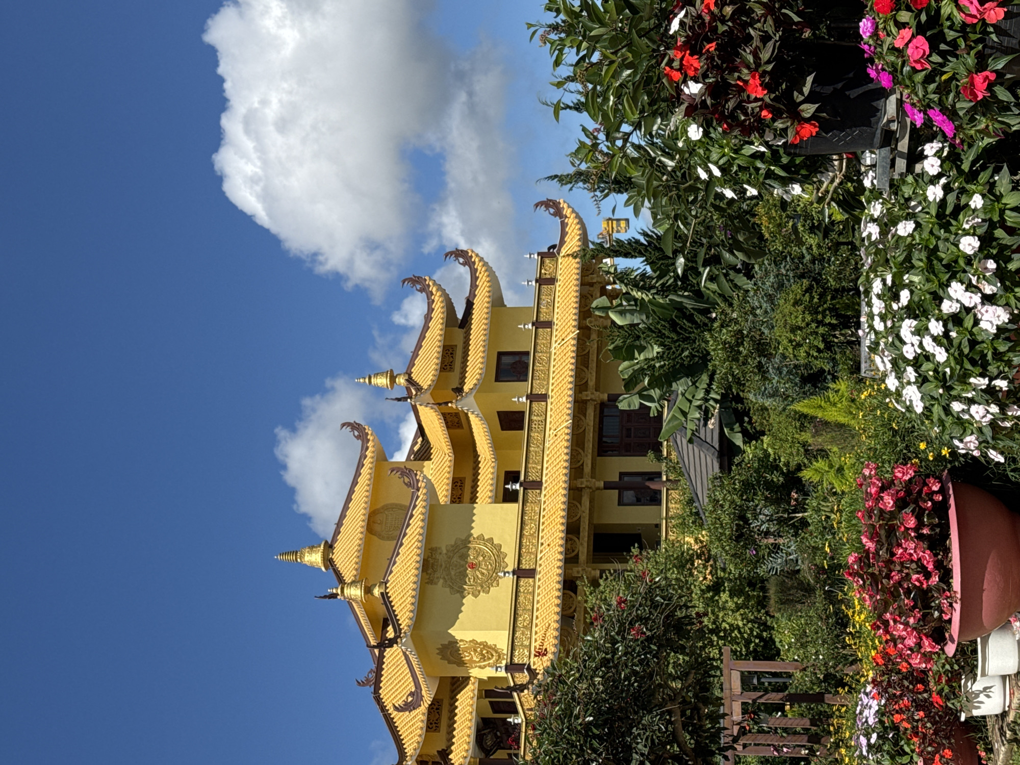 The image shows a multi-tiered, yellow Buddhist temple in Da Lat, Vietnam. The temple's architecture features curved rooflines with ornamental detailing. The sky behind the temple is blue with white clouds. The foreground includes green foliage with a variety of flowers in planters. A wooden fence is also visible.