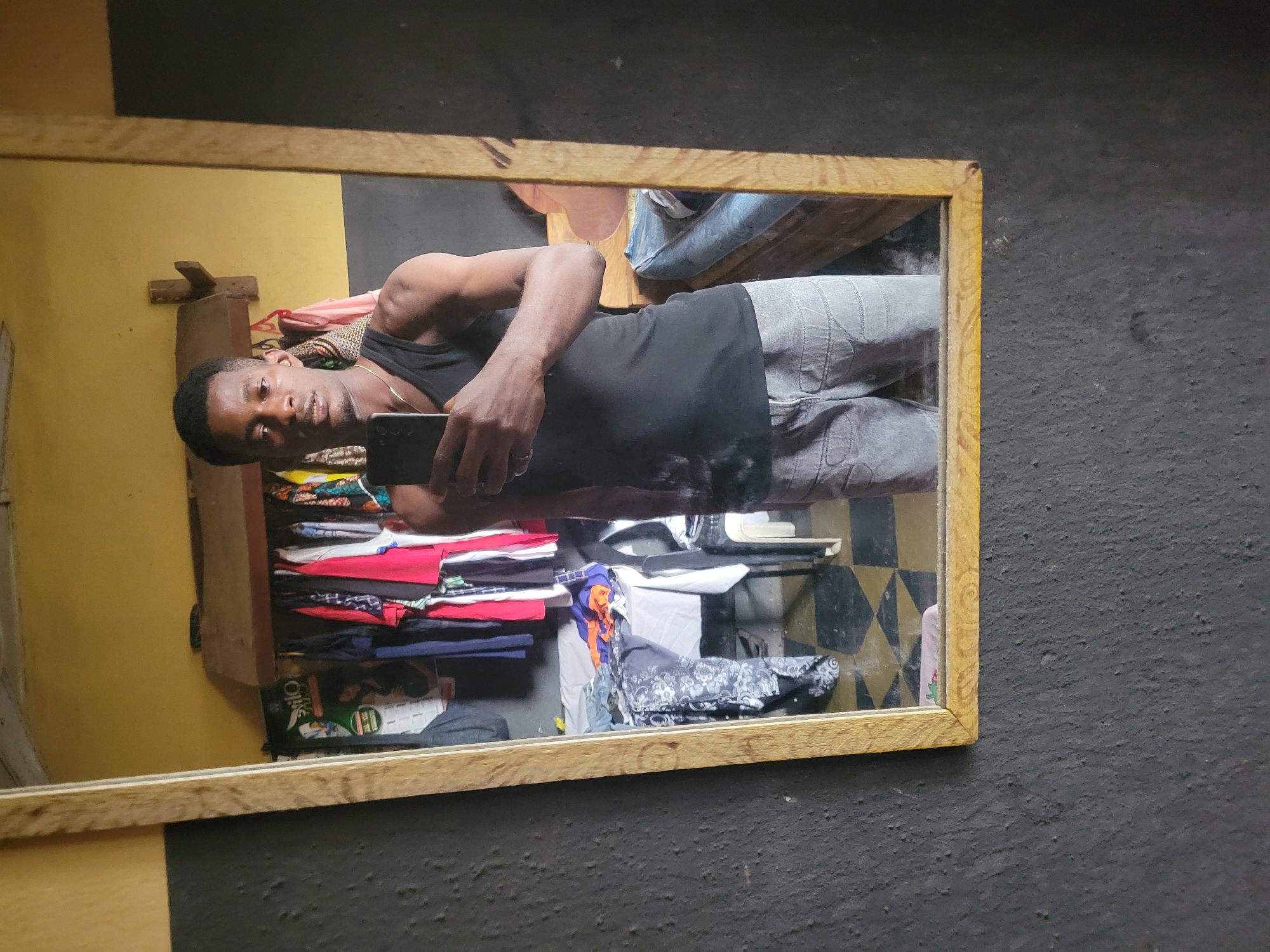 A full-body selfie is taken in a bedroom, with a mirror reflecting the subject and part of the room. The subject, a young Black male, stands facing the mirror, holding a smartphone in his right hand and taking the picture. He is wearing a black tank top and grey pants. Behind him, a rack of clothes hangs on the wall, with a table covered in laundry to the left. A bed is visible in the background, with a blue mattress. The walls of the room are painted yellow and black, with a dark, textured finish on the black sections. A wooden-framed mirror is the central element of the scene. The floor has a checkerboard pattern of black and yellow tiles.