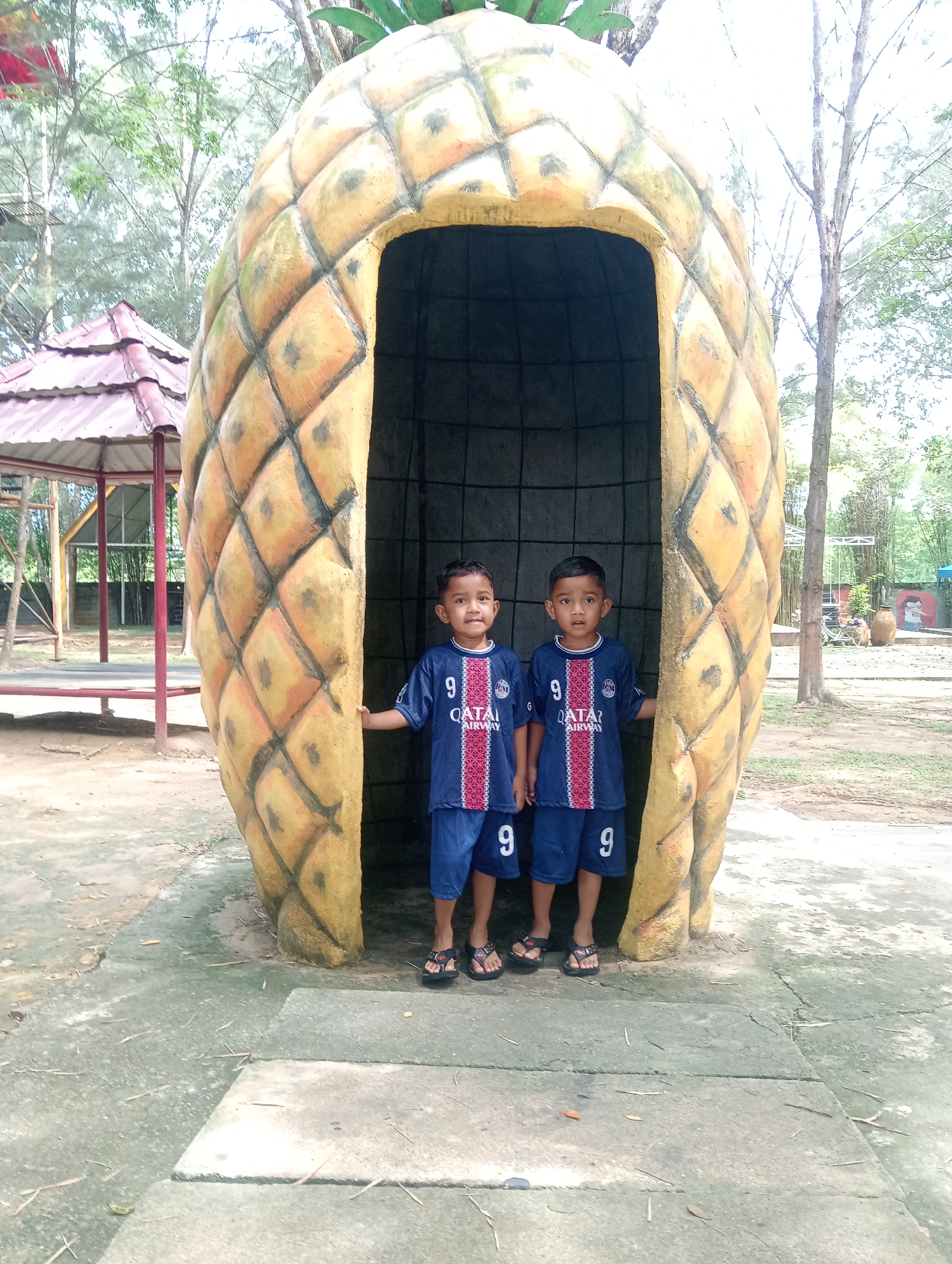 Two young boys wearing matching Paris Saint-Germain soccer jerseys and shorts stand inside a large, hollow pineapple structure. The boys are positioned side-by-side, their hands lightly touching the interior edges of the pineapple's opening. The ground is a concrete surface with visible seams and discoloration. Behind the pineapple, a small gazebo-like structure with a red roof is partially visible on the left. Trees and other greenery populate the background, with some trees appearing bare. This photo was taken in Lhokseumawe, Indonesia.