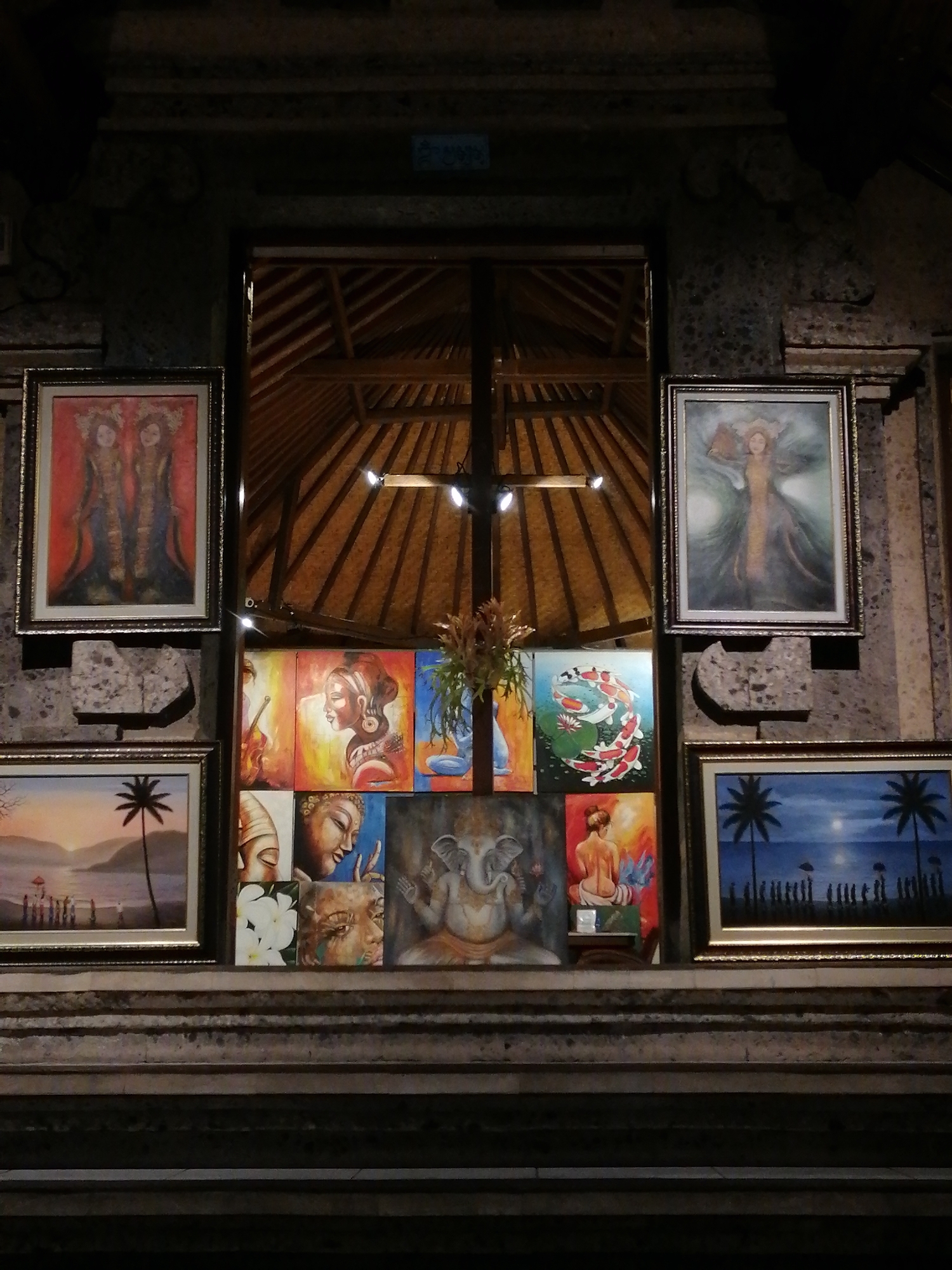 The image displays a collection of paintings arranged on stone steps in front of a building. The building features a thatched roof and stone carvings. The paintings are diverse in subject matter, including portraits of people, Buddhist imagery like Ganesha and Buddha, a scene with koi fish, and a sunset landscape with palm trees and figures. Two larger paintings are hung on the wall on either side of the entrance. The overall impression is of an art display, possibly in a market or gallery setting, located in the Ubud District of Indonesia.