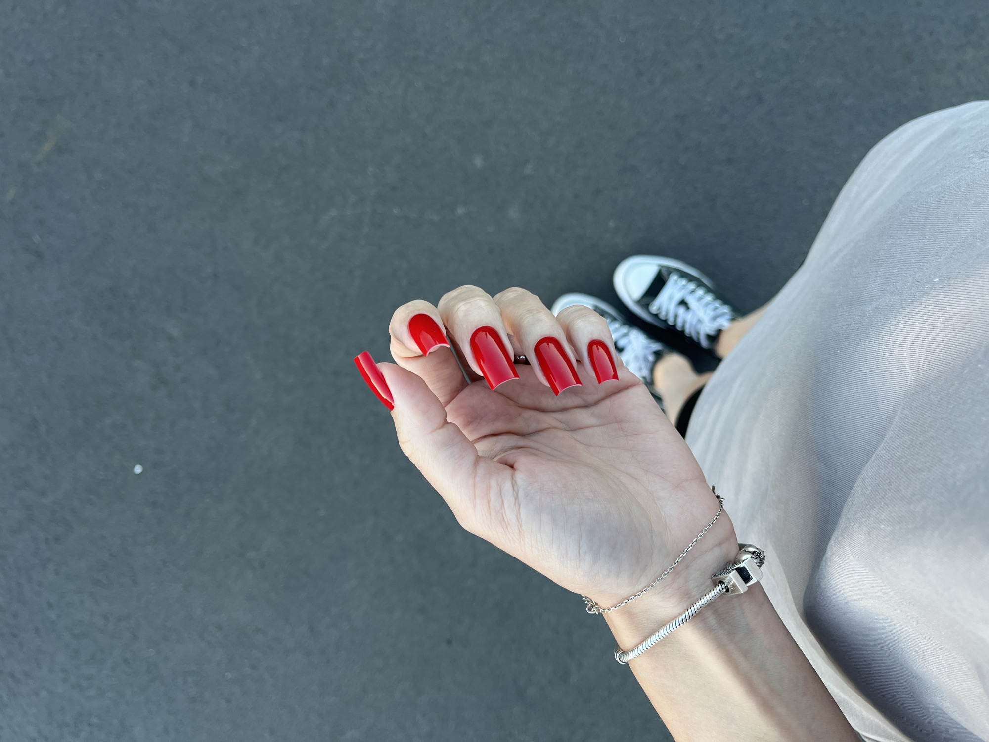 The image shows a close-up of a person's left hand with long, red, stiletto-shaped fingernails. The hand is held over a dark, asphalt-like surface, likely a road or sidewalk. In the background, partially visible, are a pair of black Converse sneakers with white laces. To the right of the hand, a portion of a light-colored, possibly beige or grey, skirt or dress is visible, suggesting the person is wearing this garment. A silver bracelet with a charm is worn on the wrist of the hand with the red nails. The lighting and shadows suggest it is daytime, possibly outdoors. The scene is casual and everyday, with no specific event indicated. The location context provided is Nizhny Novgorod, Russia, but there are no overt visual cues in the image to confirm this specific location. There is no visible text in the image.