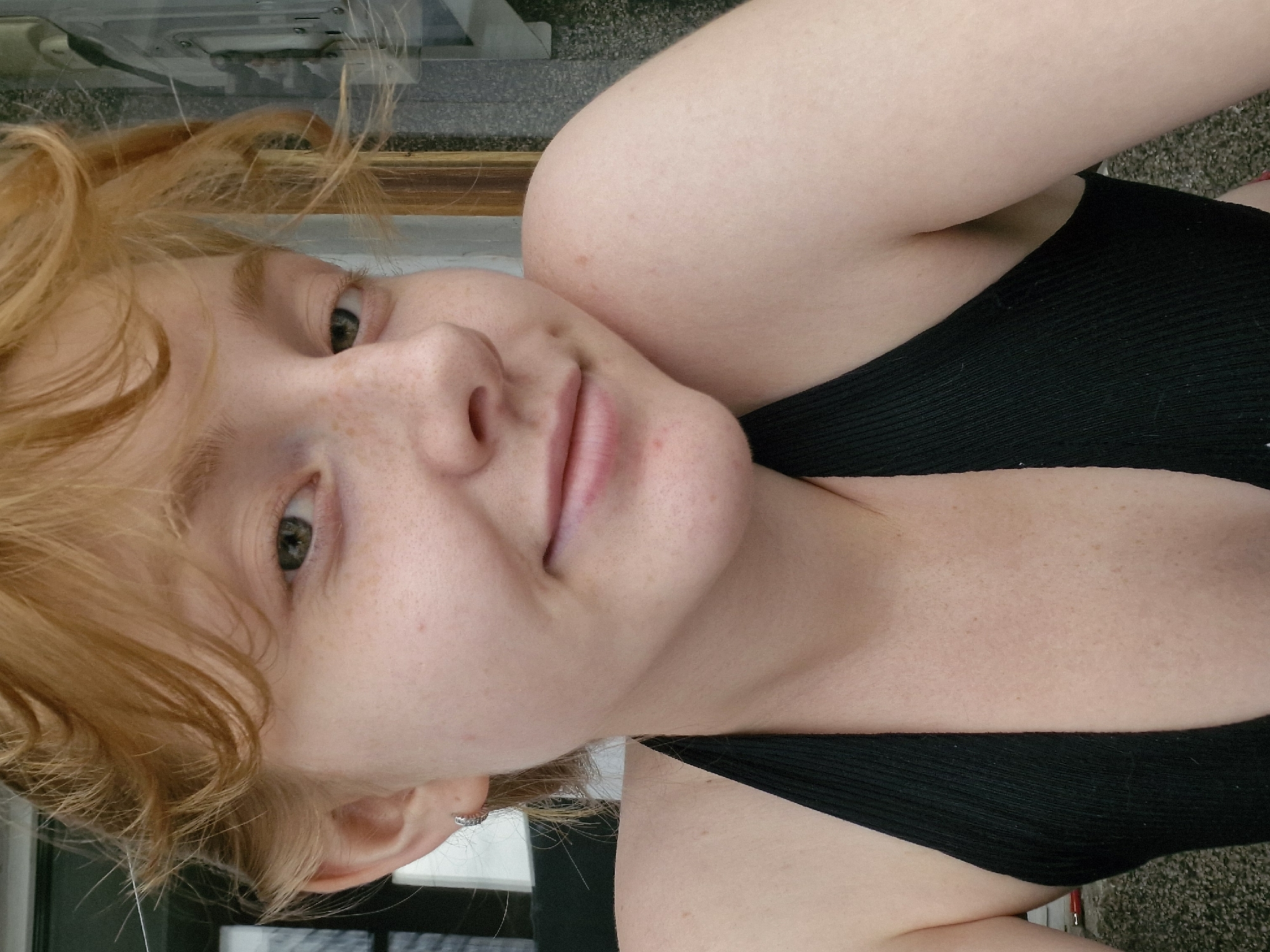 The image is a close-up, shoulder-up selfie of a young person with fair skin, freckles, and short, wavy auburn hair. They are wearing a black halter top. The background is out of focus and appears to be an outdoor setting, possibly a porch or patio, with a building structure visible. There is no identifiable information within the image that would place it in Yekaterinburg, Russia.
