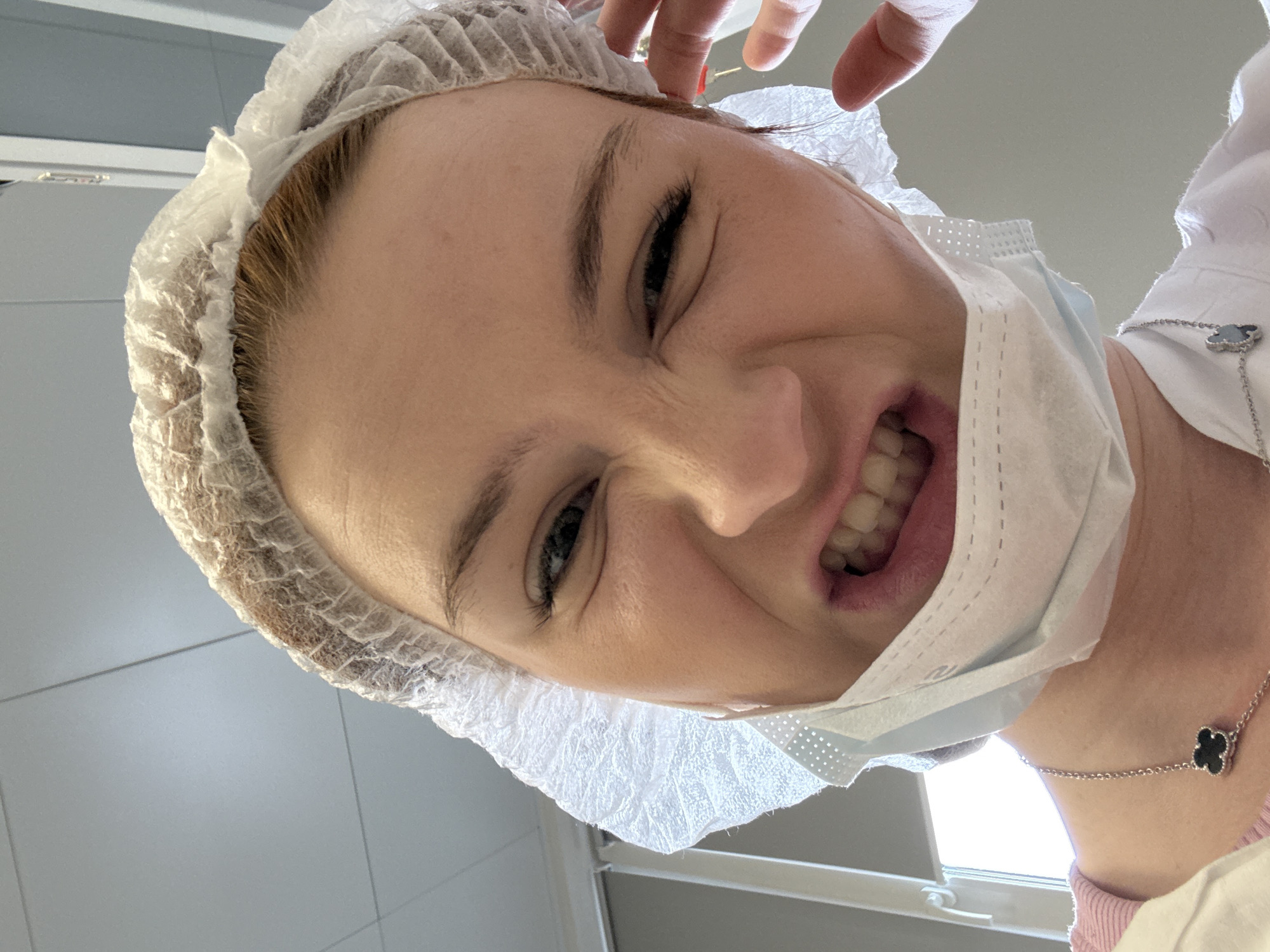 A low-angle view captures a woman in a surgical cap and mask, her face contorted in a grimace. She wears a necklace with a four-leaf clover pendant. A hand is visible near her head. The background is a sterile-looking environment, possibly a medical or dental setting. The presence of a door suggests an interior space. No indication of the city of Saint Petersburg, Russia, is discernible.