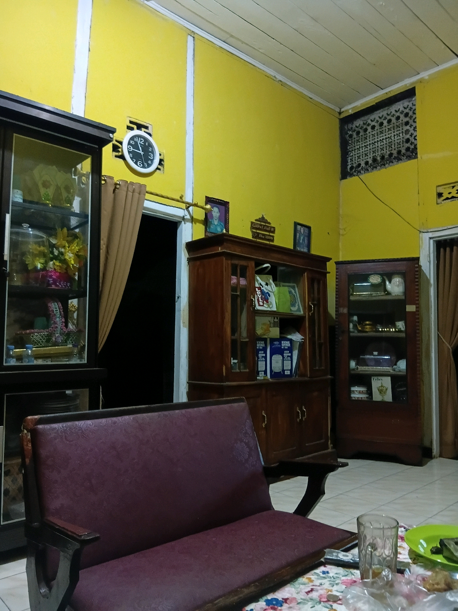 The image depicts an indoor scene, likely a living room or dining area, in what appears to be a home in Terbanggi Marga, Indonesia. The walls are painted a vibrant yellow. In the foreground, a dark-colored sofa with a patterned burgundy upholstery is visible, its right armrest and back showing signs of wear. A low table in front of the sofa is covered with a floral tablecloth. On the table, a clear glass with a light-colored liquid, a remote control, a small plate with food, and a plastic bag containing items are present.
The background features several pieces of wooden furniture. To the left, a tall, dark-colored glass-fronted cabinet displays various items, including flowers and what looks like decorative objects. To the right of this, a large wooden cabinet with glass doors showcases more items, including what appears to be a teapot and serving dishes. Further to the right, another tall glass-fronted wooden cabinet contains various objects on its shelves.
On the yellow wall, a circular clock with black numerals and hands is prominently displayed. To the right of the clock, a framed photograph of a person in uniform is hung. Above the furniture, decorative elements like a patterned ventilation grate and other framed pictures are visible. A brown curtain hangs near a doorway leading to a dark space. The ceiling is made of wooden planks. The floor appears to be tiled. The overall impression is of a lived-in domestic space, possibly reflecting an older style of interior design.