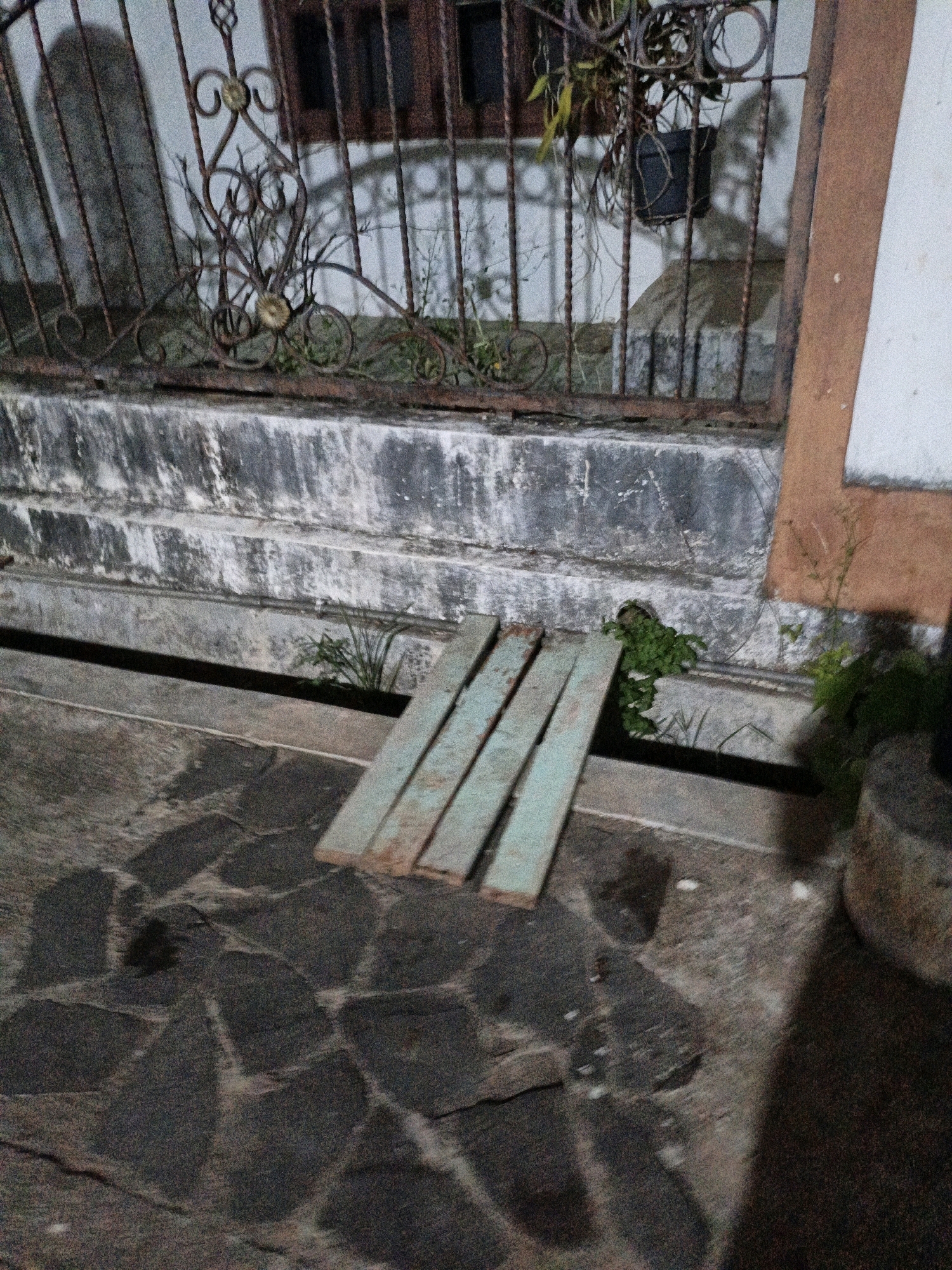 The image shows an outdoor scene at ground level, likely in Cikal, Indonesia. In the foreground, there is a paved area made of dark, irregularly shaped stones, with some lighter colored debris or markings scattered across it. Three light blue wooden planks are laid diagonally across the paving stones, extending from the bottom right towards the top left. To the left of the planks, there is a narrow, dark horizontal channel or drain. Behind this drain, a weathered concrete wall or structure runs horizontally. Further back, a rusty metal fence with decorative elements and vertical bars separates the area from a building. The fence has intricate scrollwork and some gold-colored embellishments. Behind the fence, a building with dark windows and a pot of greenery hanging from a plant is visible. Greenery is also growing around the base of the concrete wall and in the drain. The overall lighting suggests it is either dawn, dusk, or nighttime.