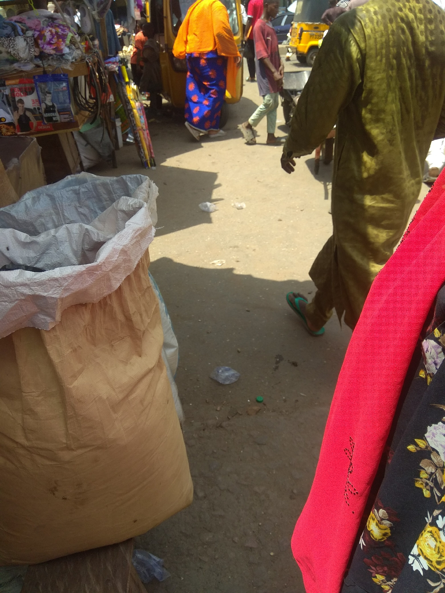 The image depicts a street scene in Jalingo, Nigeria, likely a market area. In the foreground, a large tan burlap sack is partially visible, with a white plastic liner. To the right of the sack, a person wearing a vibrant red fabric is partially visible, with a dark fabric patterned with flowers underneath. In the background, another person is seen from the back, wearing a long, olive-green garment. This individual is walking away from the camera and is wearing green flip-flops. Further in the background, a yellow tricycle is parked, and other people are present, including one wearing an orange top and a blue and orange patterned skirt. The ground appears to be a dirt or asphalt surface, with some debris scattered around. The overall impression is of a busy outdoor marketplace.