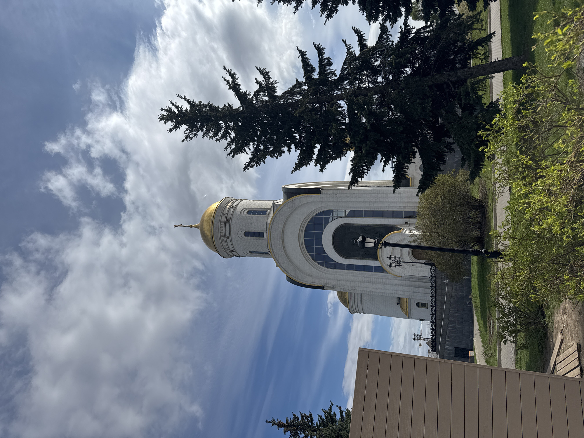 The image shows a view of the Church of St. George in Victory Park, Moscow, Russia. The white stone church features a prominent golden dome and is partially obscured by a conifer tree and other vegetation. The sky is visible with a mix of blue sky and white clouds. A lamppost and a wooden fence are also in view.