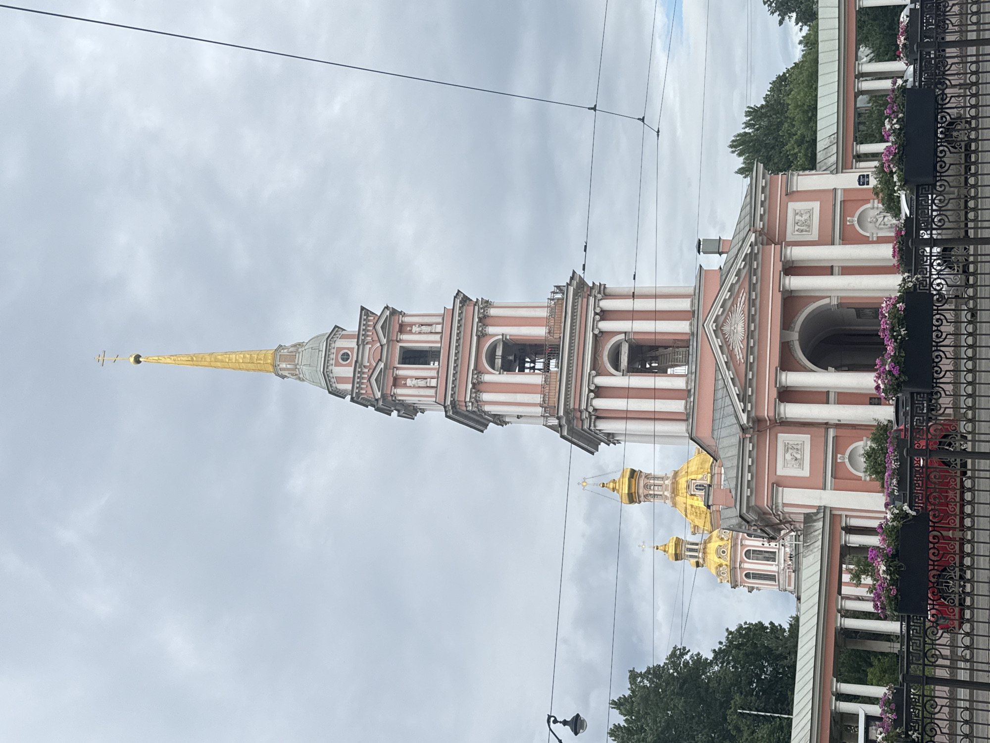 The image captures a view of the Smolny Cathedral in Saint Petersburg, Russia, on an overcast day. The prominent feature is the cathedral's tall, pink and white bell tower, topped with a golden spire and a cross. Below the spire, the tower has multiple levels with arched openings where bells are visible. The main body of the cathedral, also predominantly pink, features classical architectural elements such as columns, pediments, and decorative reliefs. Two golden domes of the cathedral are visible behind the bell tower. In the foreground, a black metal fence encloses a walkway lined with black planters filled with pink and white flowers. A red car is partially visible behind the fence, along with other vehicles. Overhead, power lines crisscross the sky. Lush green trees frame the sides of the scene. The overall impression is one of grand religious architecture under a somber sky.