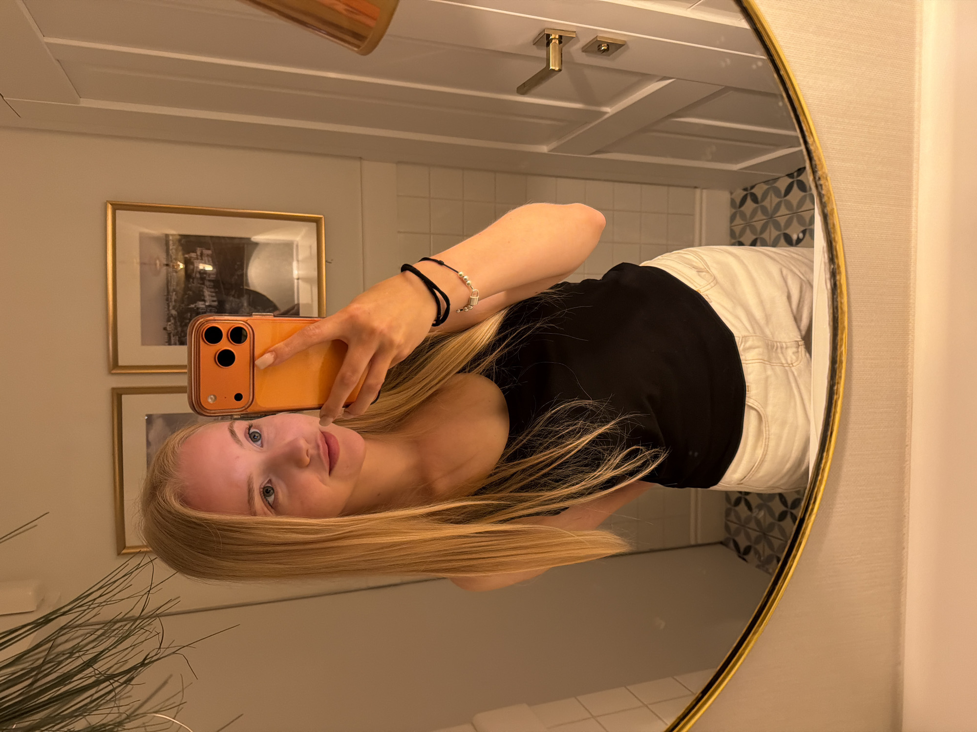 A blonde woman is reflected in a gold-framed mirror. She is holding an orange iPhone. She is wearing a black shirt and white pants. The background of the reflection shows a bathroom with tiled walls and framed pictures. There are plants near the mirror. The location is the city of Unknown, Unknown.