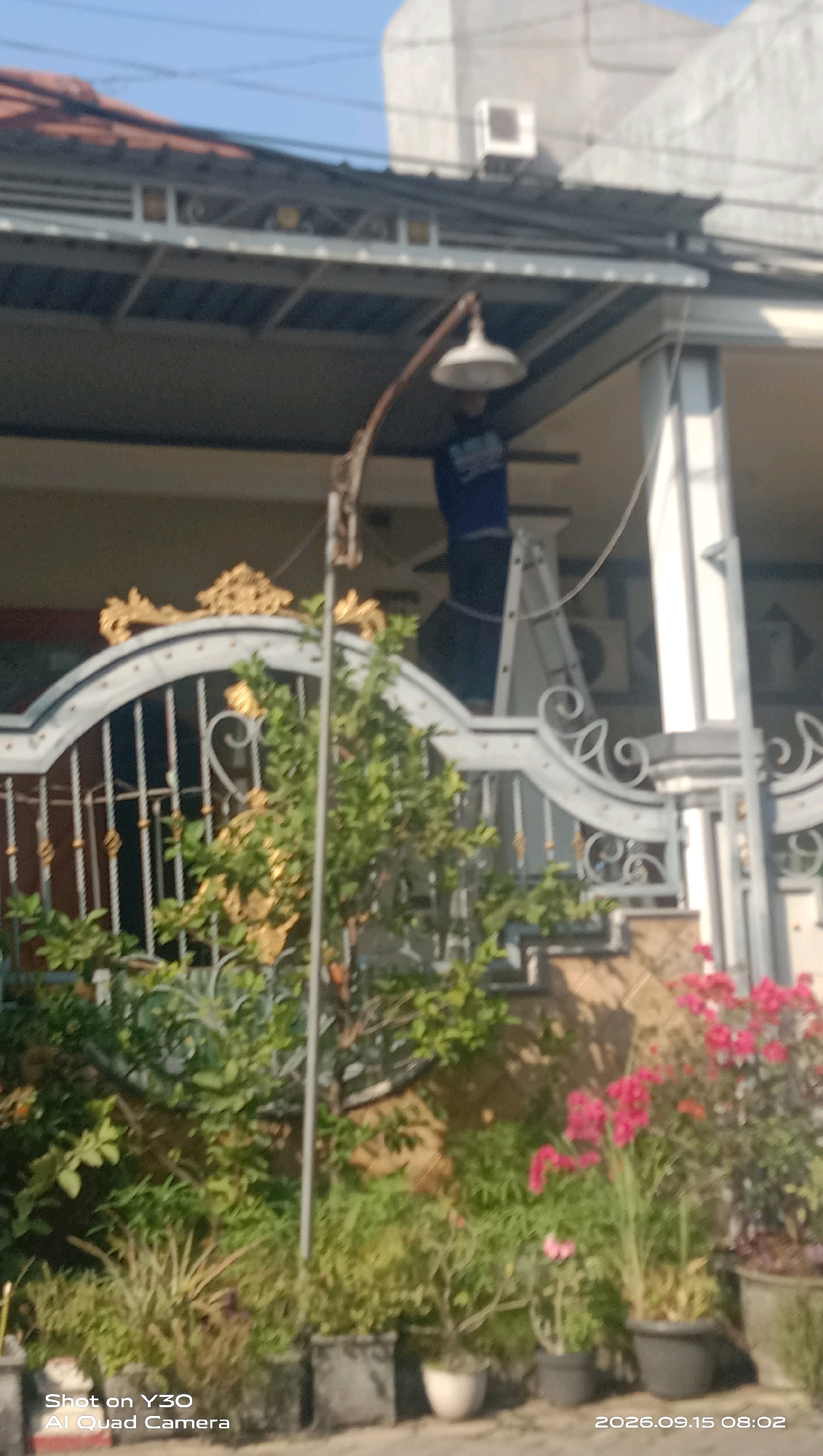 A person in a blue shirt is standing on a ladder near a house in Sekarkurung, Indonesia. The person appears to be working on a light fixture attached to the eaves of the house. In front of the house, there is a decorative metal gate with gold accents. Lush green plants and pink flowers in pots are visible in front of the gate and extending towards the right side of the image. The sky is clear and blue. The bottom left corner of the image contains the text "Shot on Y30 Al Quad Camera" and the bottom right corner has the date and time "2026.09.15 08:02".